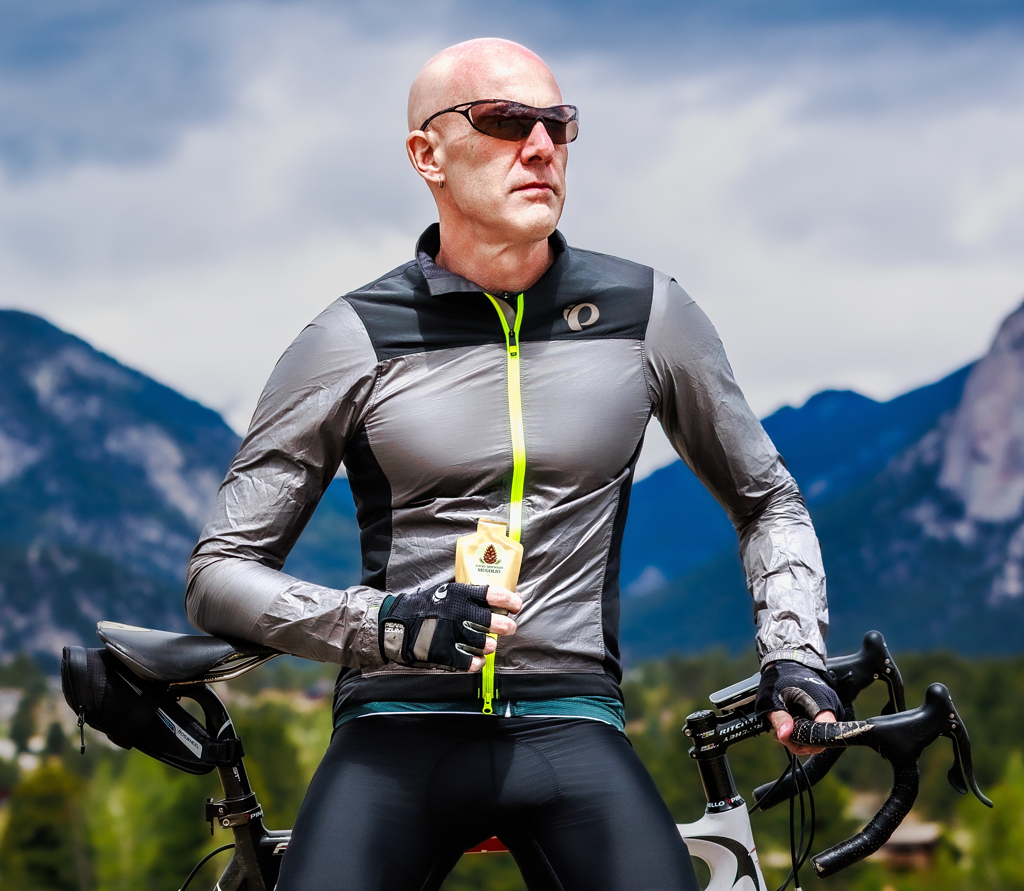 Bald man wearing sunglasses and cycling gear, holding a yellow device, with mountains and sky in background.