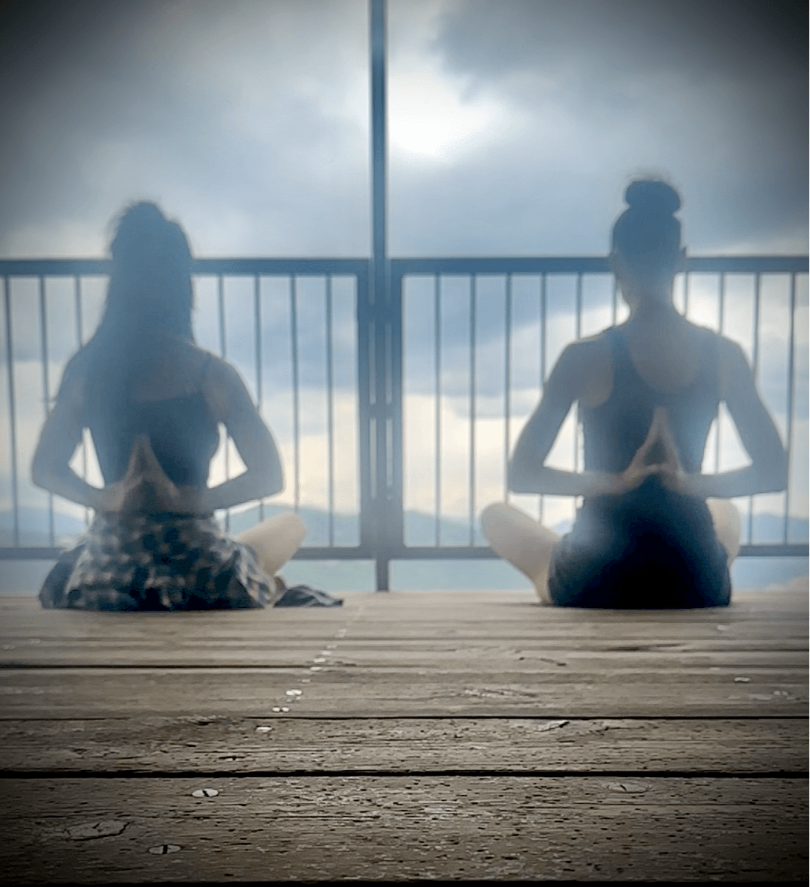 Two people sit cross-legged on a wooden deck, facing a railing with cloudy sky in the background, practicing meditation or yoga.