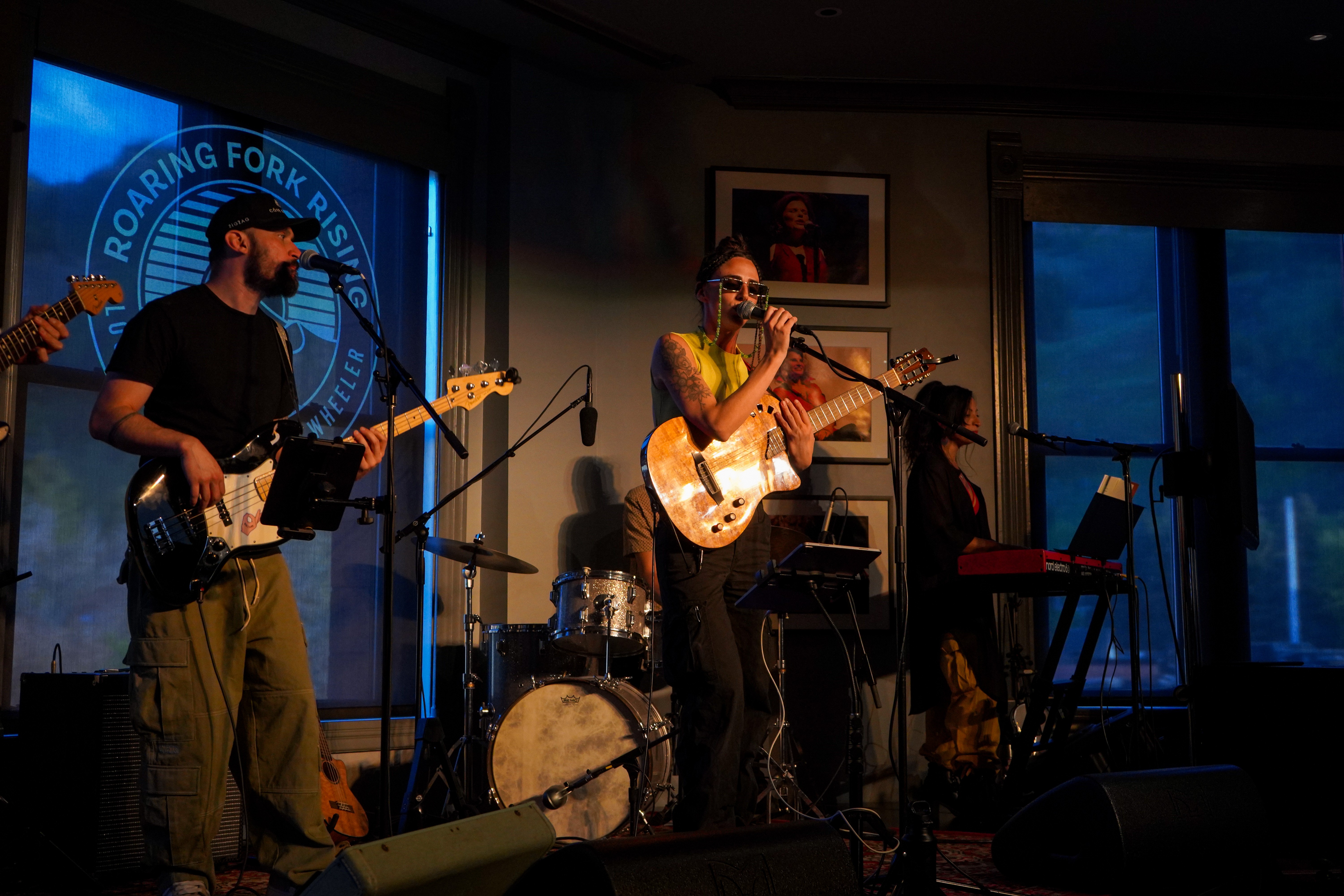 Band performs live on stage with instruments, a drummer, and a keyboardist, in a dimly lit room with a window view.