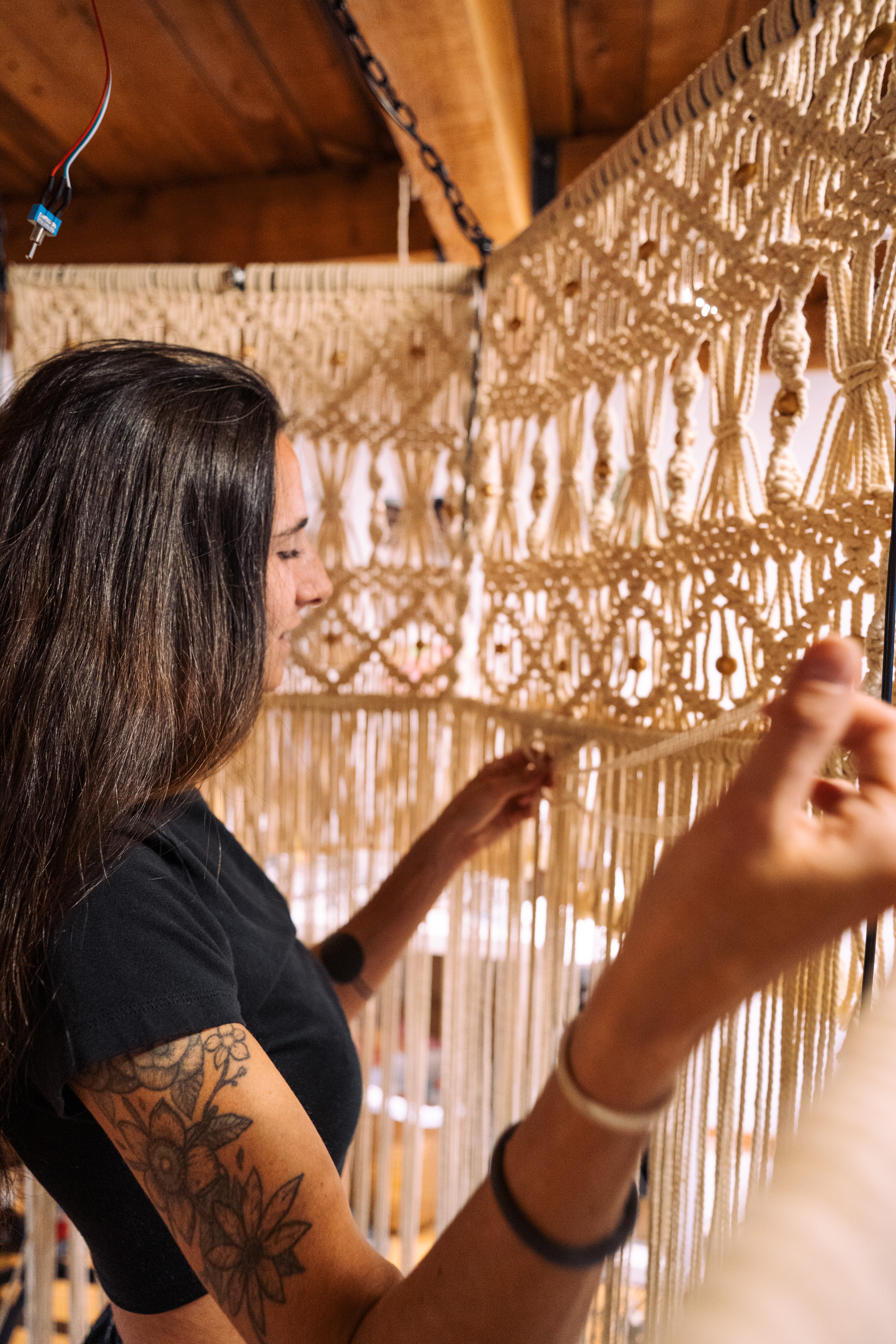 Woman with dark hair and tattoo on arm, examining a woven wall hanging, in a wooden room.