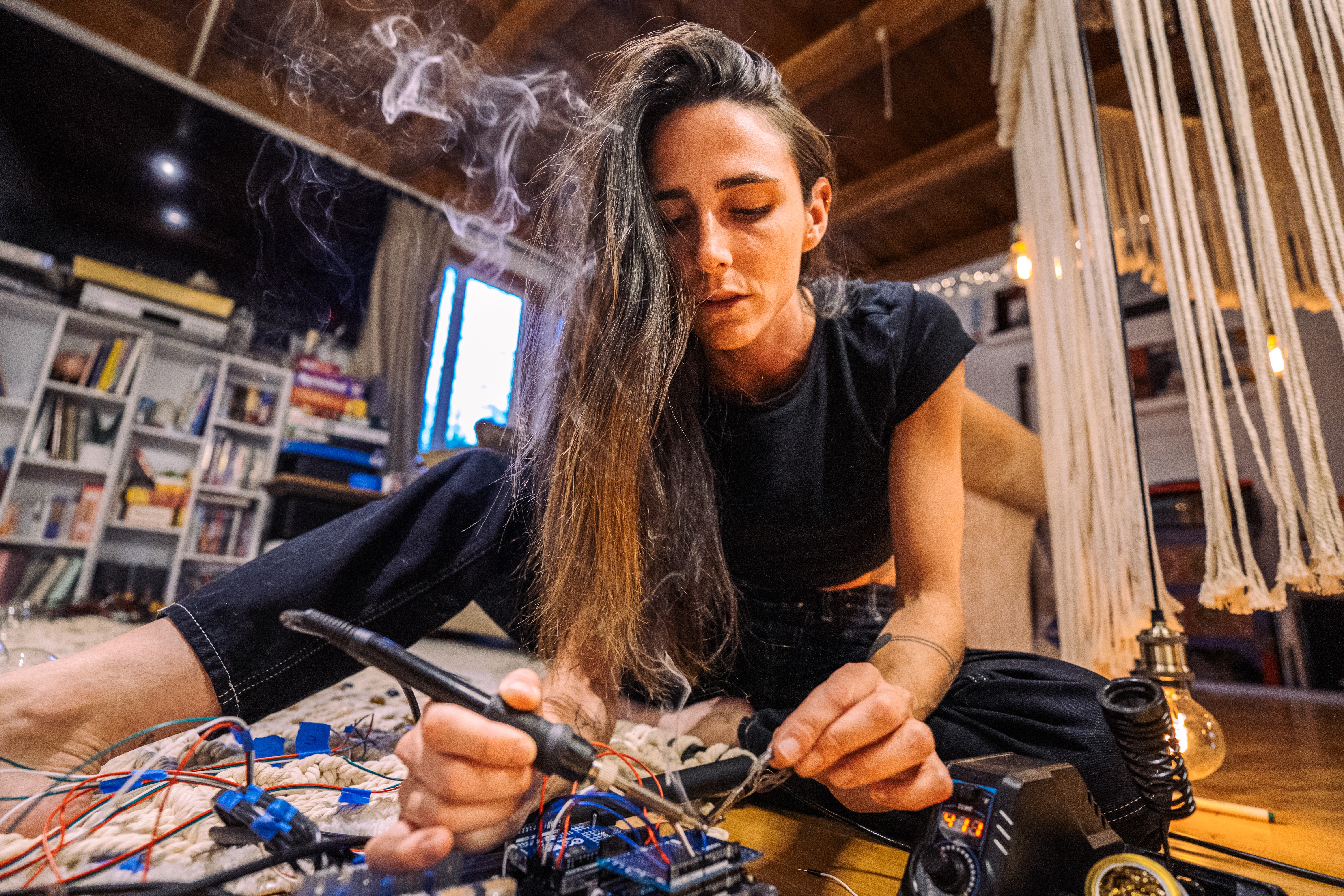 Woman with long hair working with electronic equipment indoors, surrounded by books and furniture.