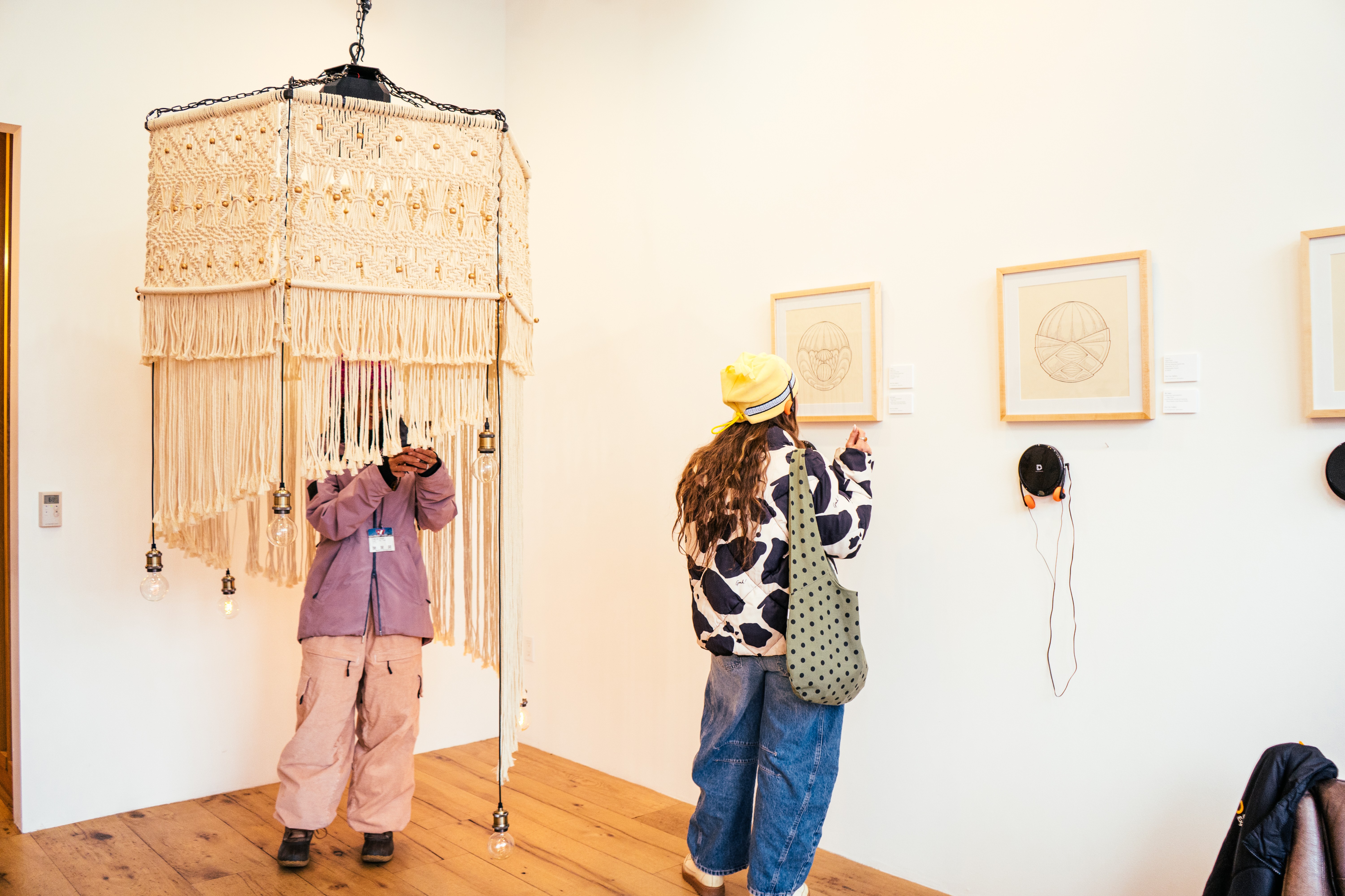 Two women stand in an art gallery, looking at framed artworks on the wall, with a hanging decorative piece nearby.