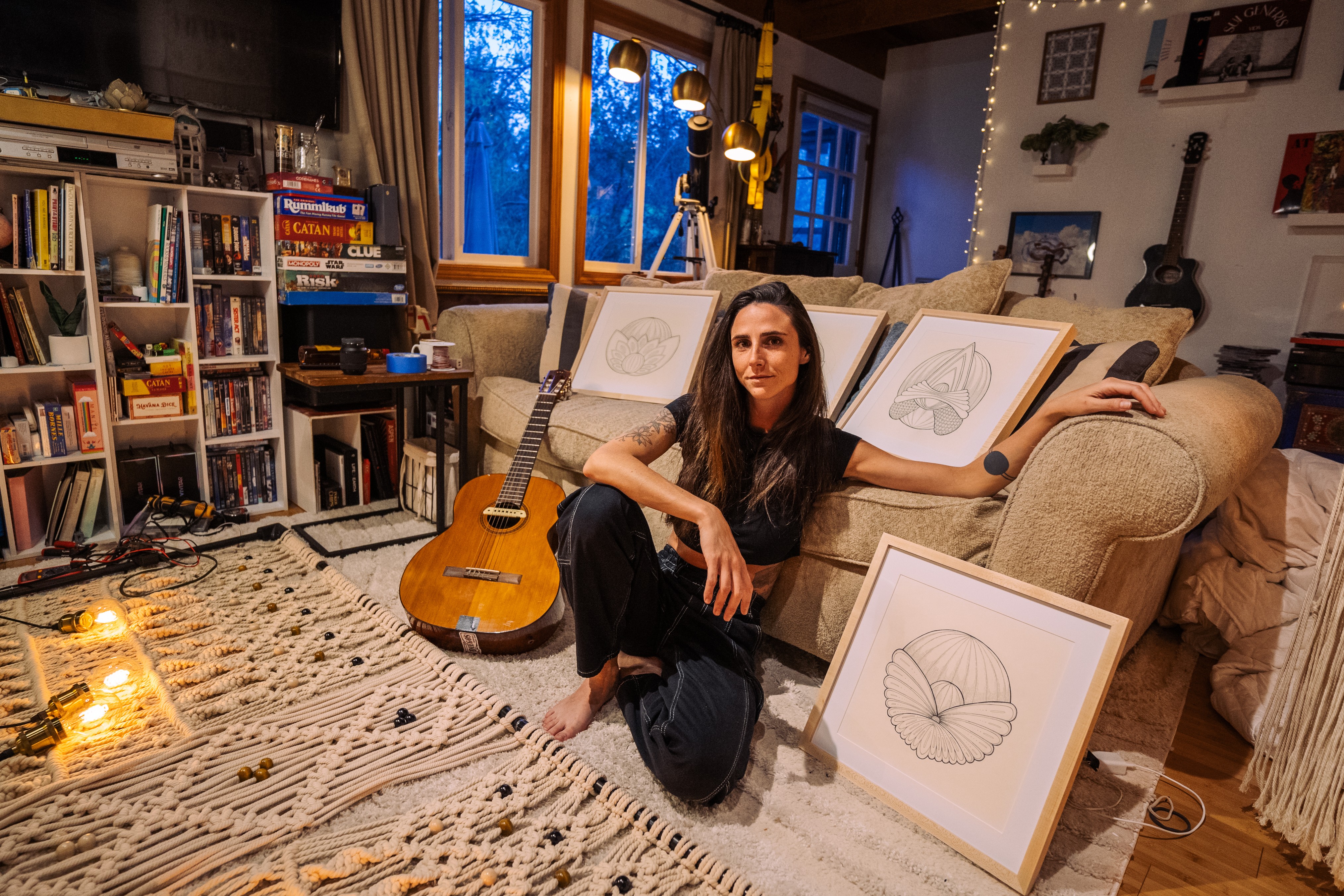 Woman sitting on a sofa in a cozy room with bookshelves, framed artwork, and a guitar nearby, during evening light.