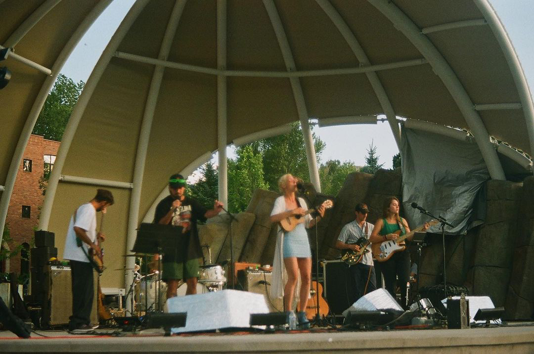 Group of six people performing on stage under a curved roof, with trees and sky in the background.