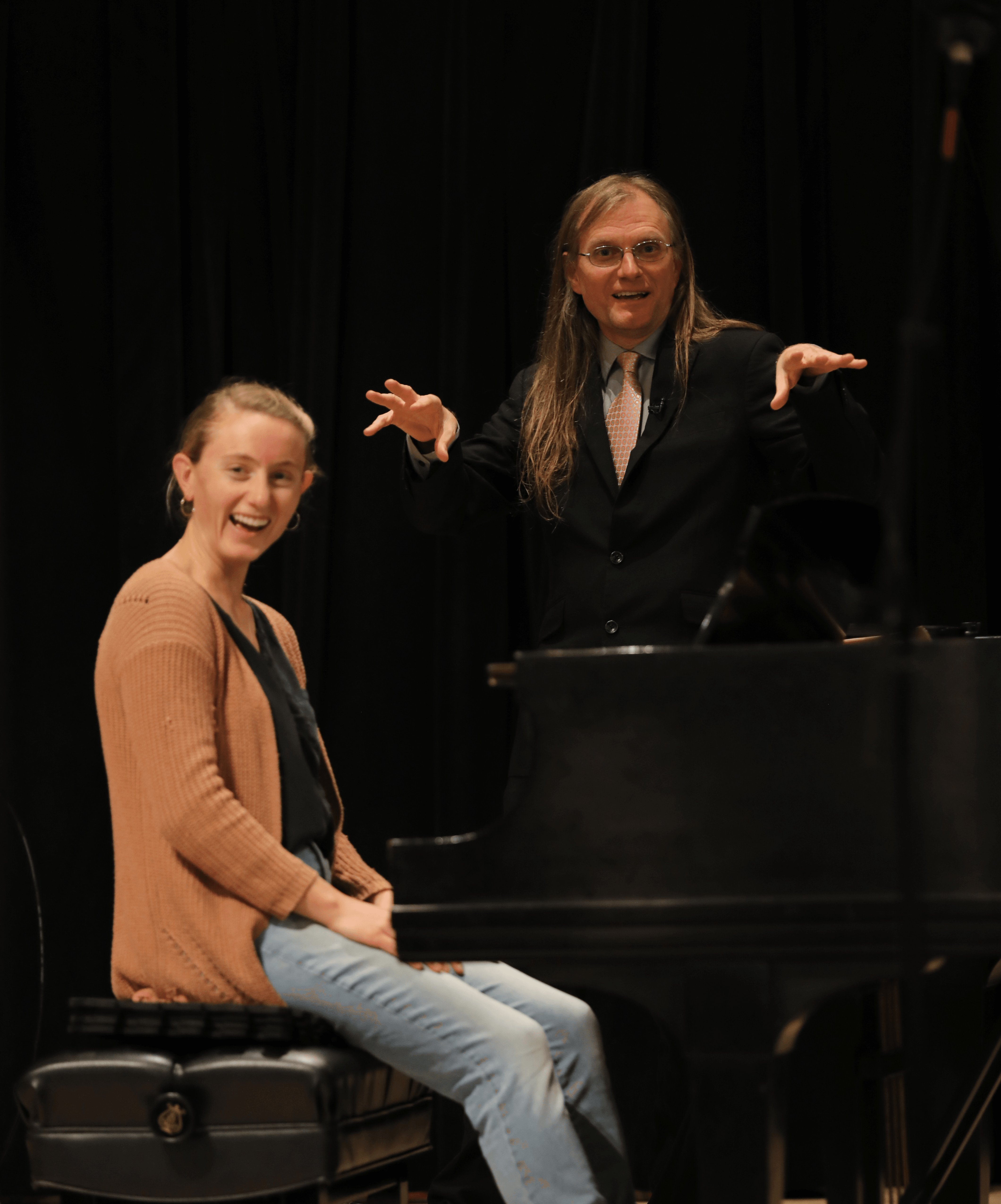 A woman with long hair and glasses stands behind a piano, gesturing with her hands, while a smiling young woman sits on a piano bench.