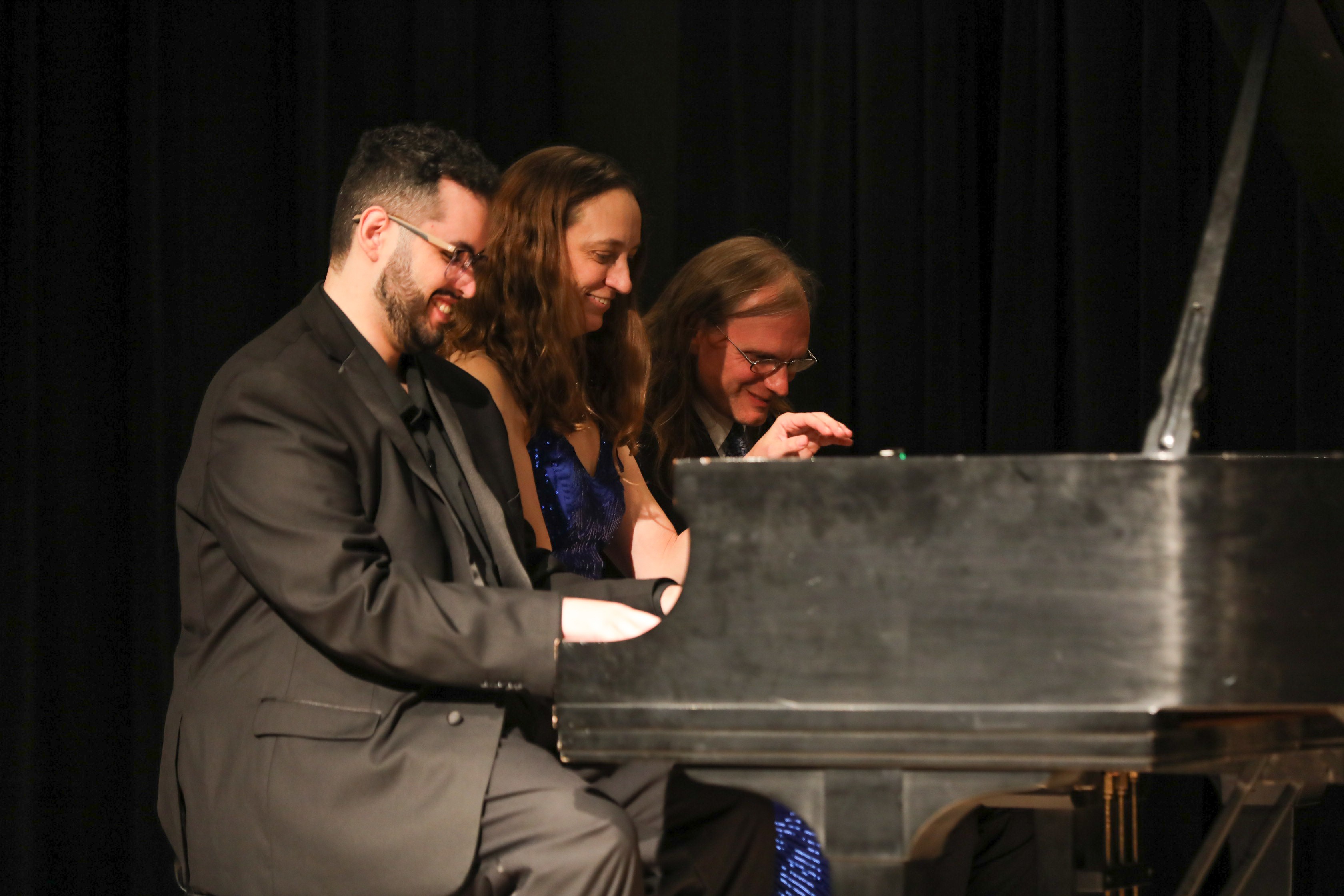 Three people sit at a piano, smiling and looking at the keys, with a dark curtain background.