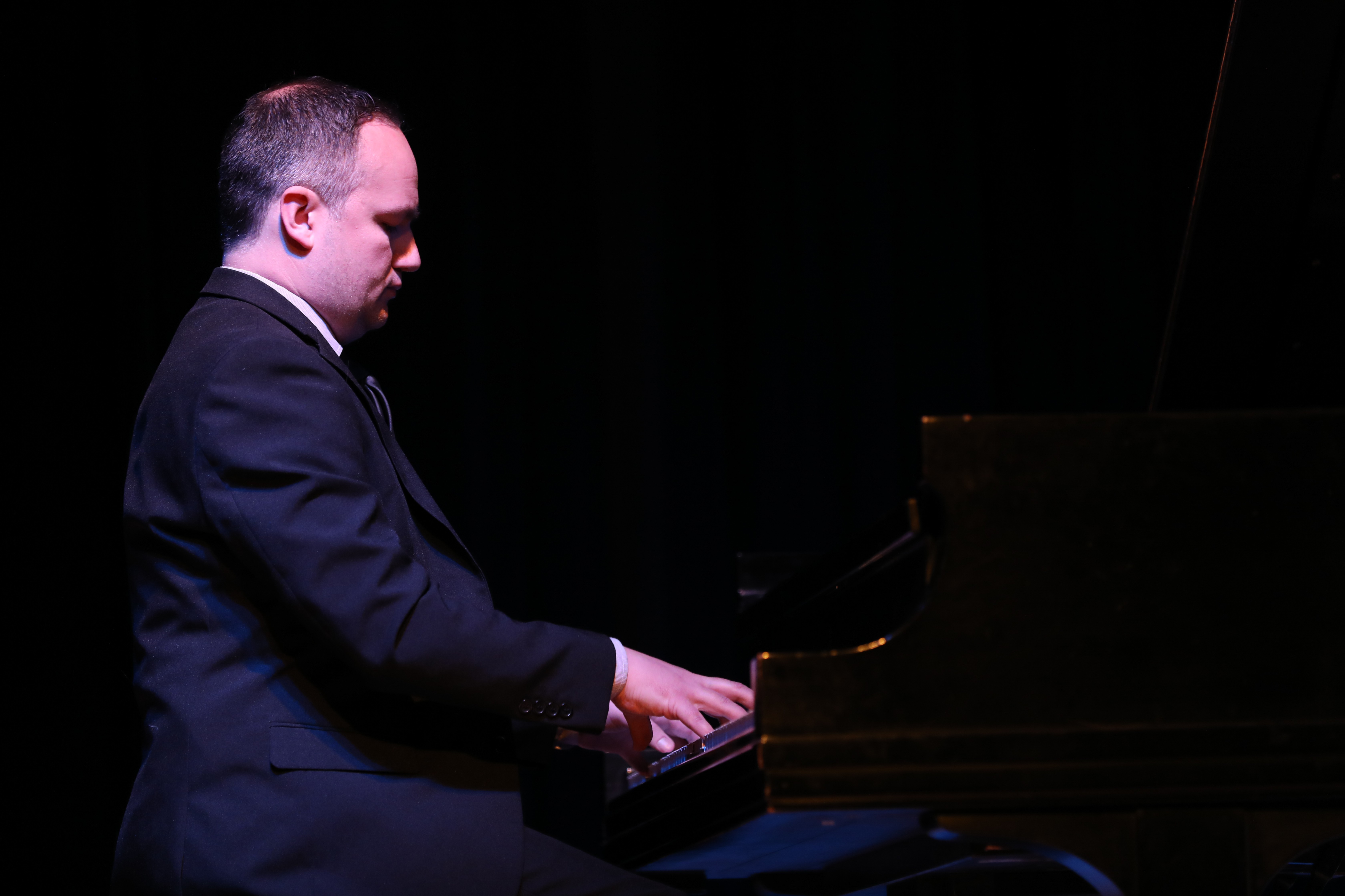 Man playing piano on stage with dark background, wearing a suit, focused on the keys.