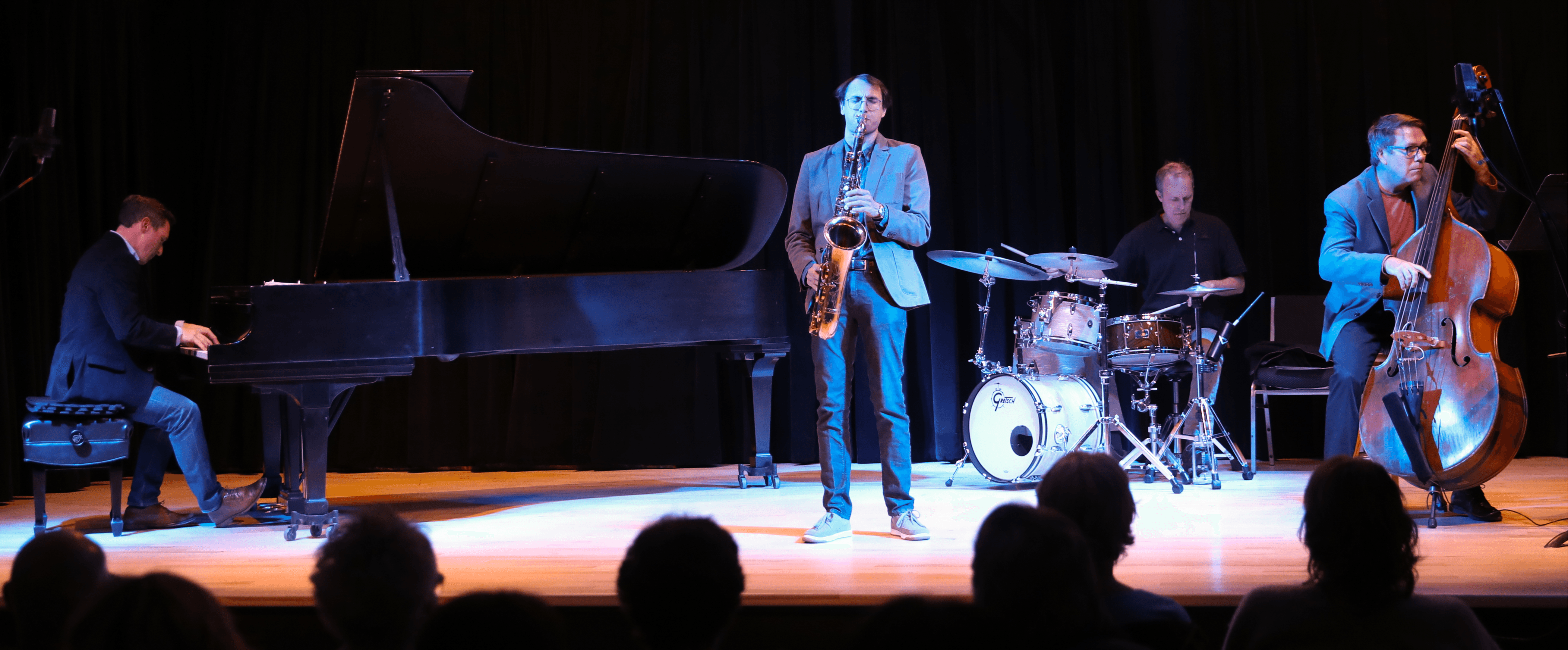 Four musicians perform on stage with a grand piano, saxophone, drums, and double bass, in front of an audience.