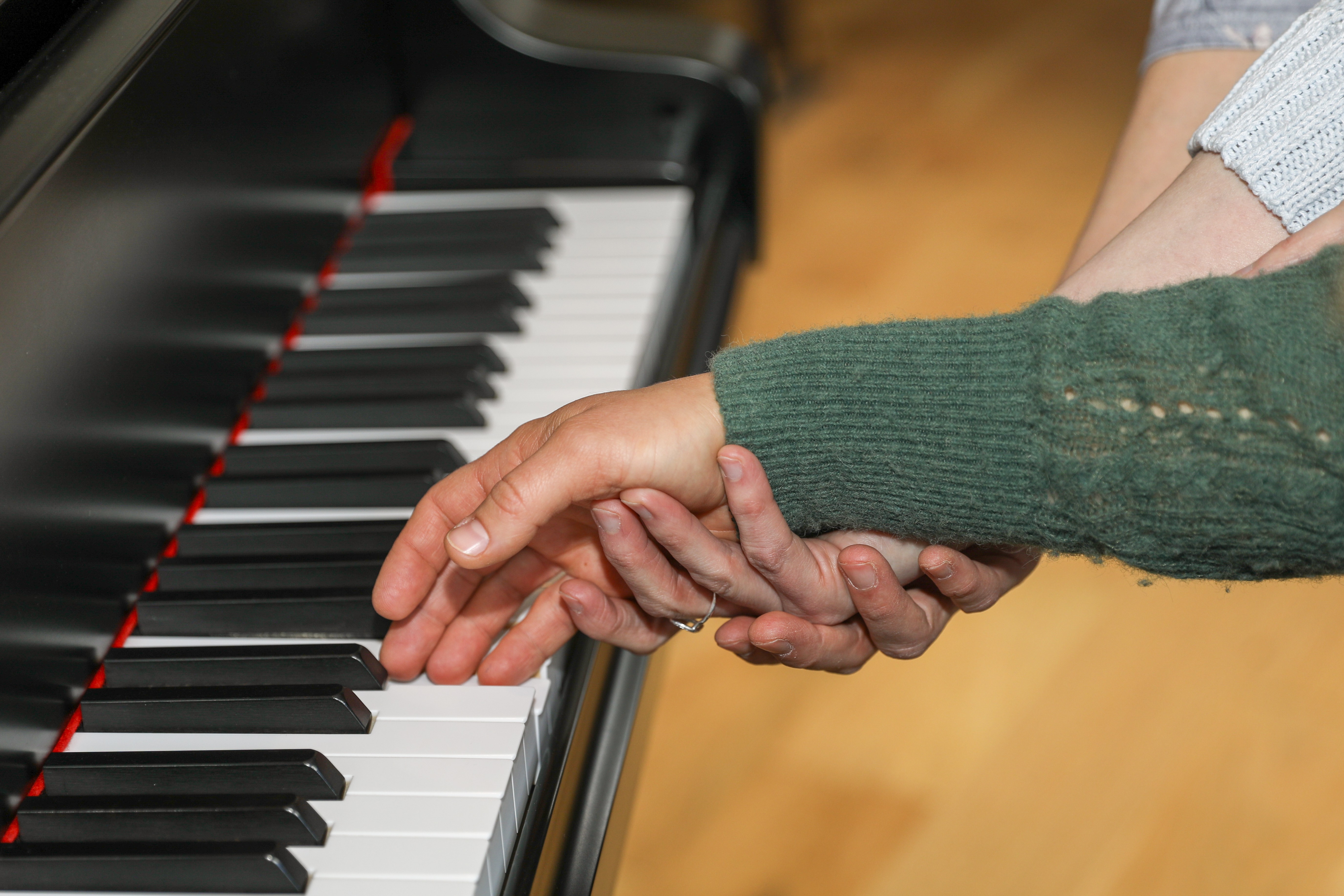 Two people hold hands above a piano keyboard, one wearing a green sweater, the other a light-colored sleeve.