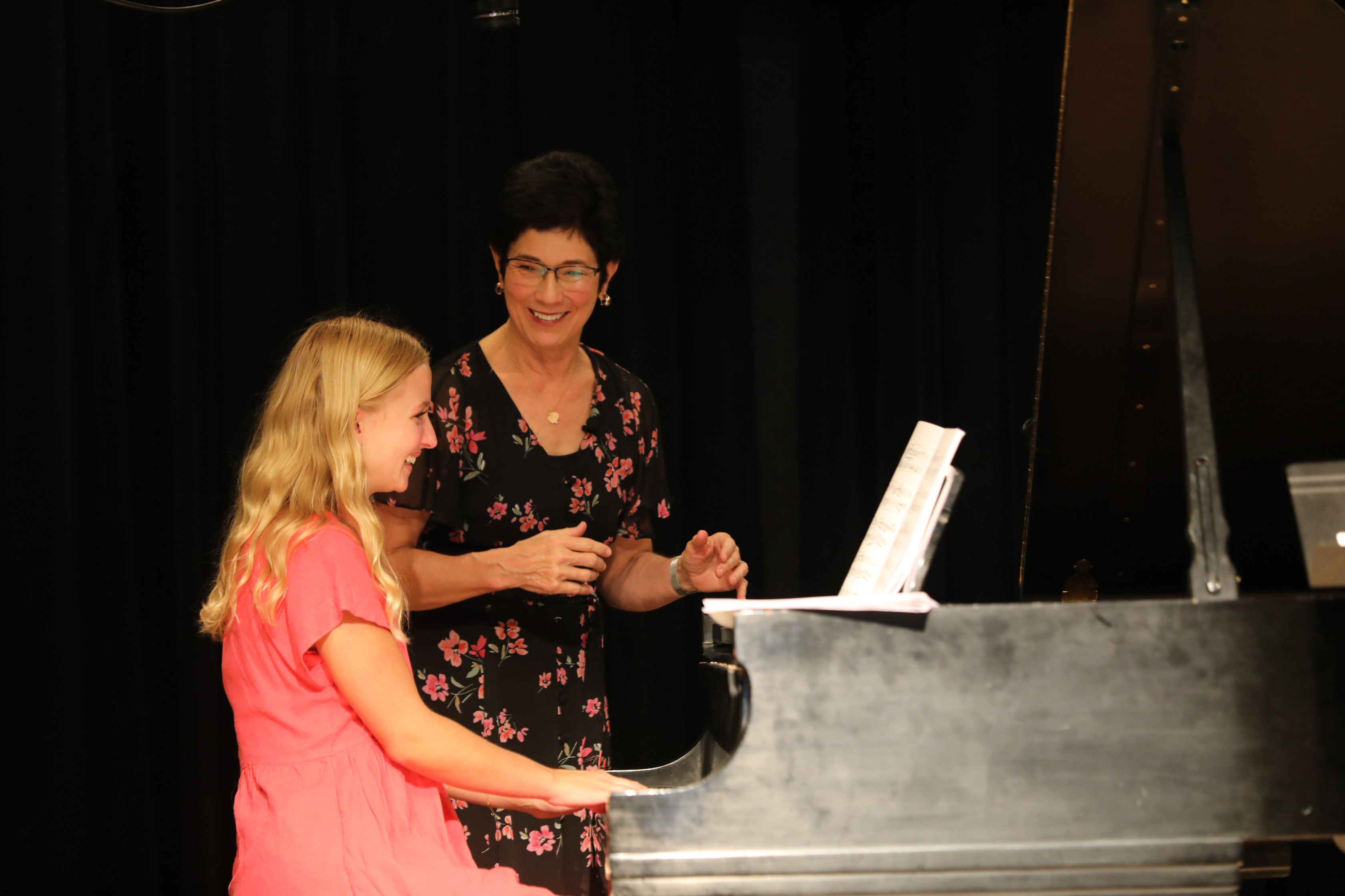 Two women near a piano, one playing and the other watching, in a dark room.