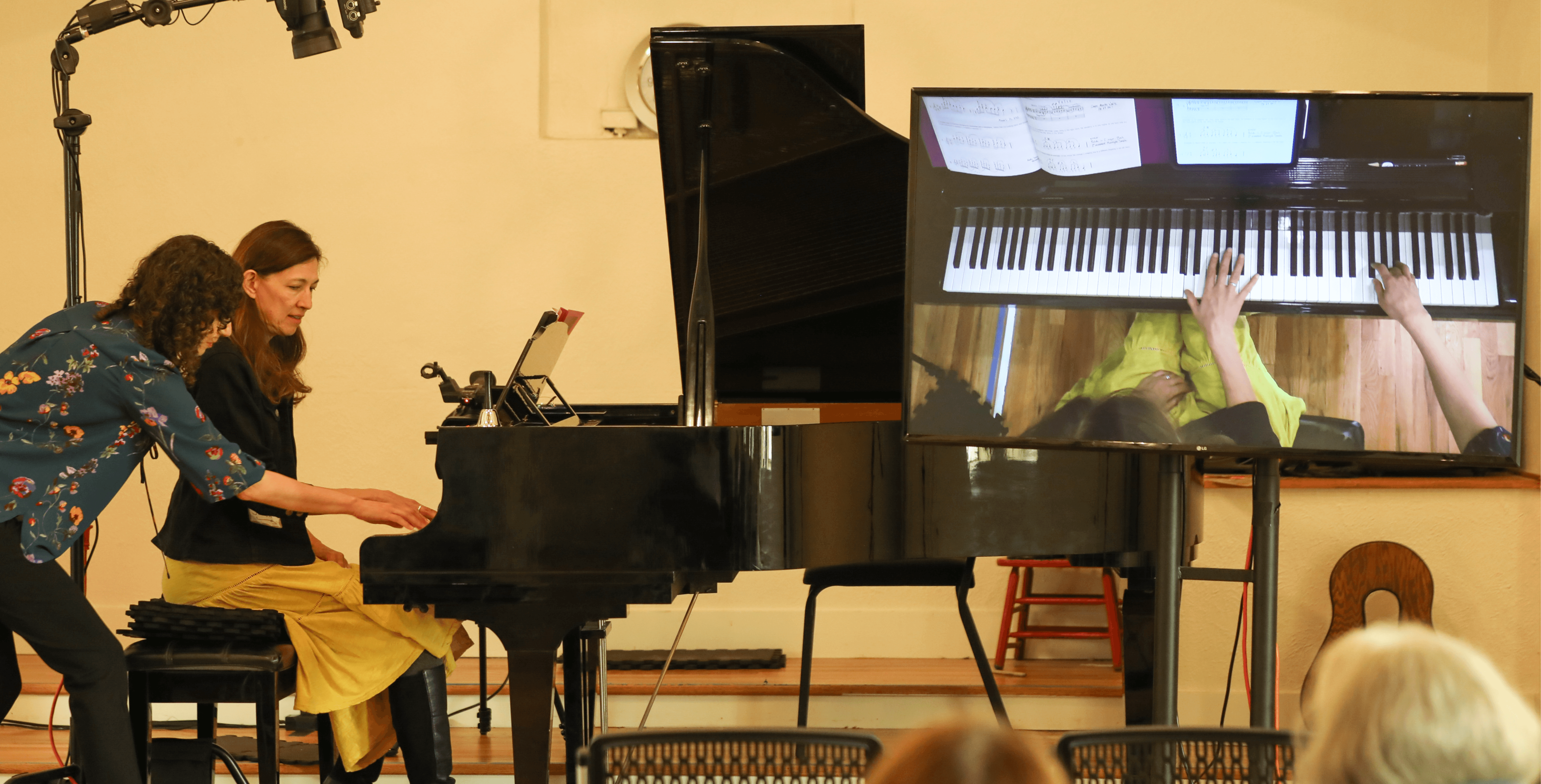 Two women play piano together, with a large screen showing a close-up of the keyboard, in a room with chairs.