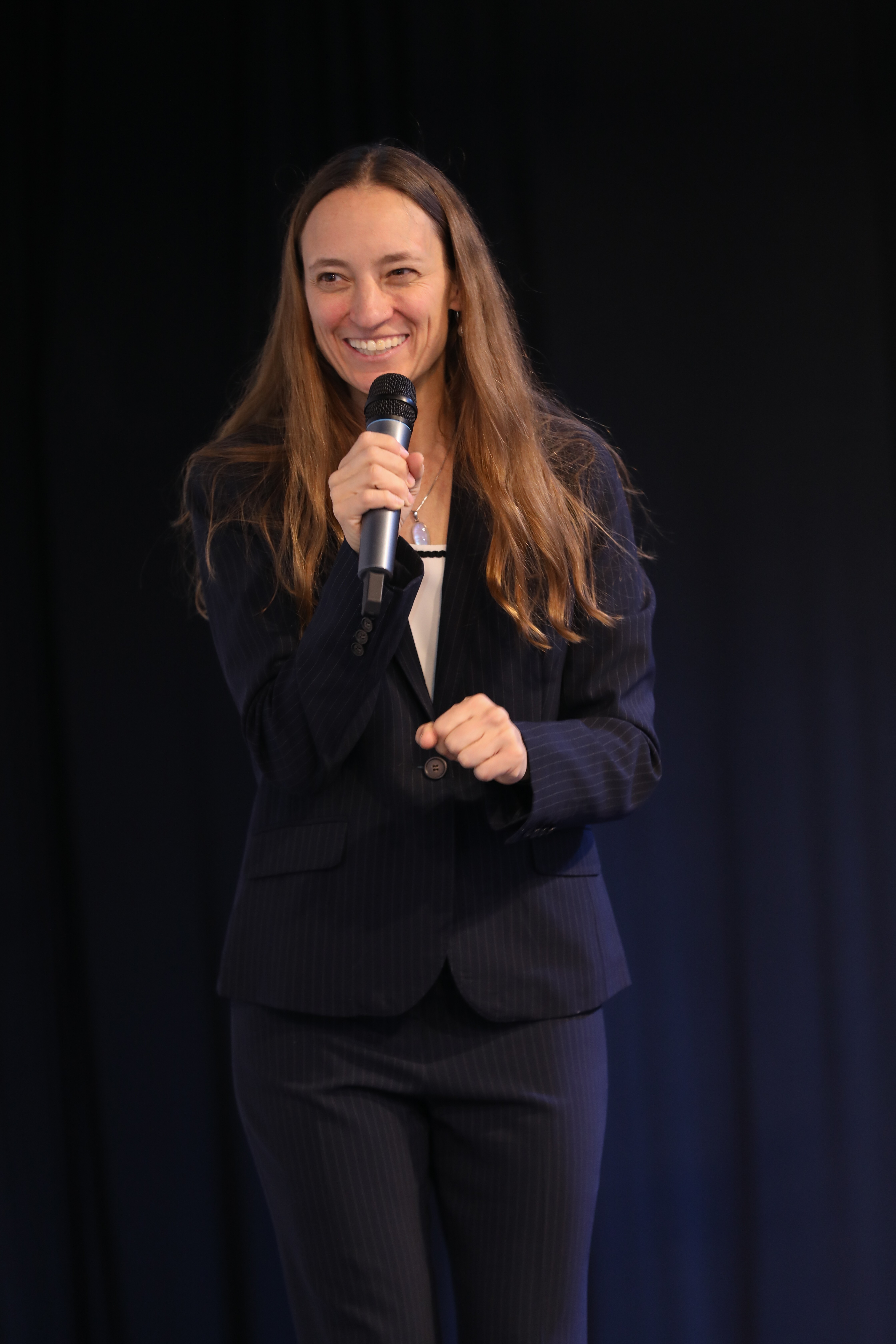 Woman smiling and speaking into a microphone on stage, wearing a dark suit, with a dark background.