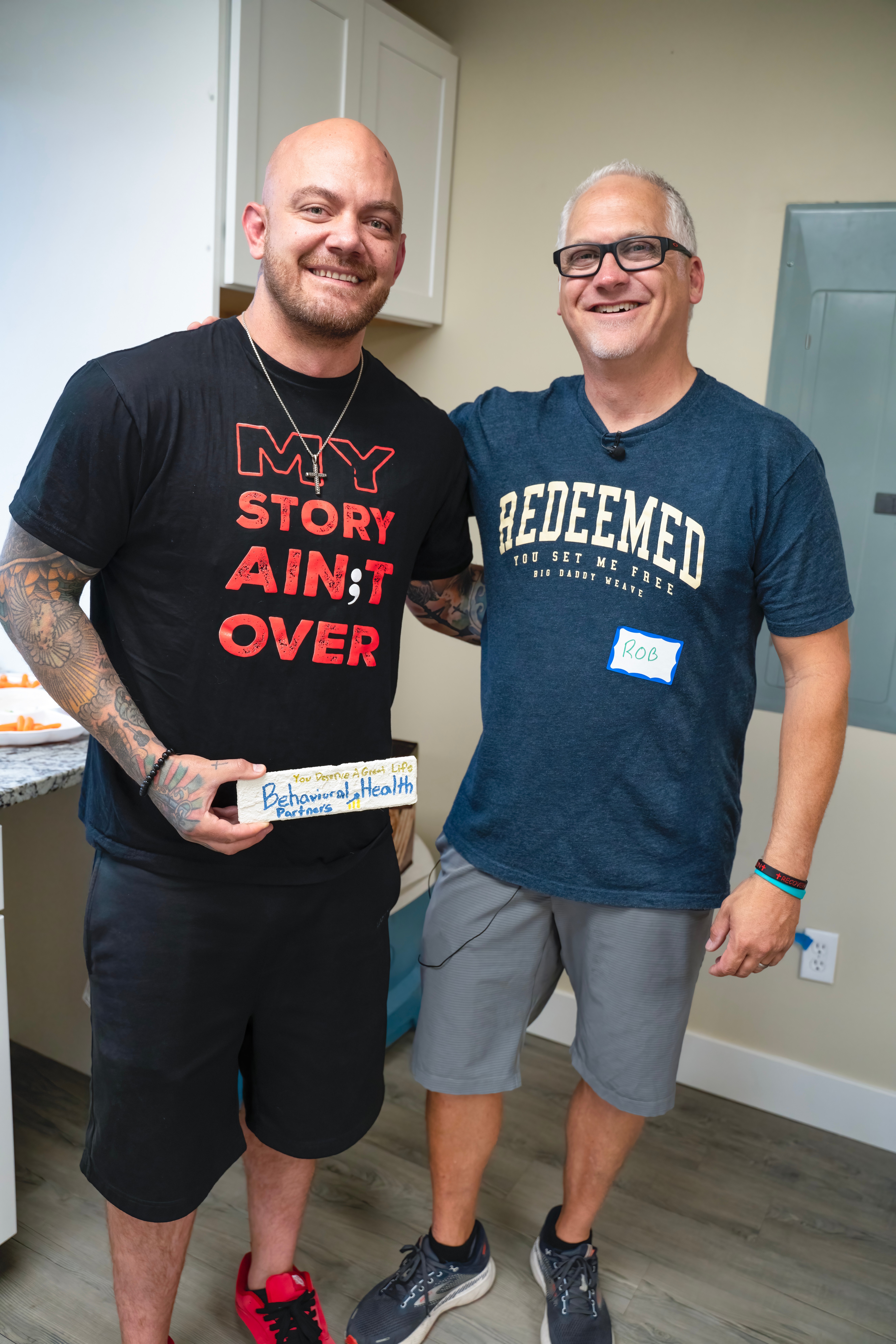 Two smiling men standing indoors, one holding a small box, the other wearing glasses and a blue T-shirt, in a room with light-colored walls.