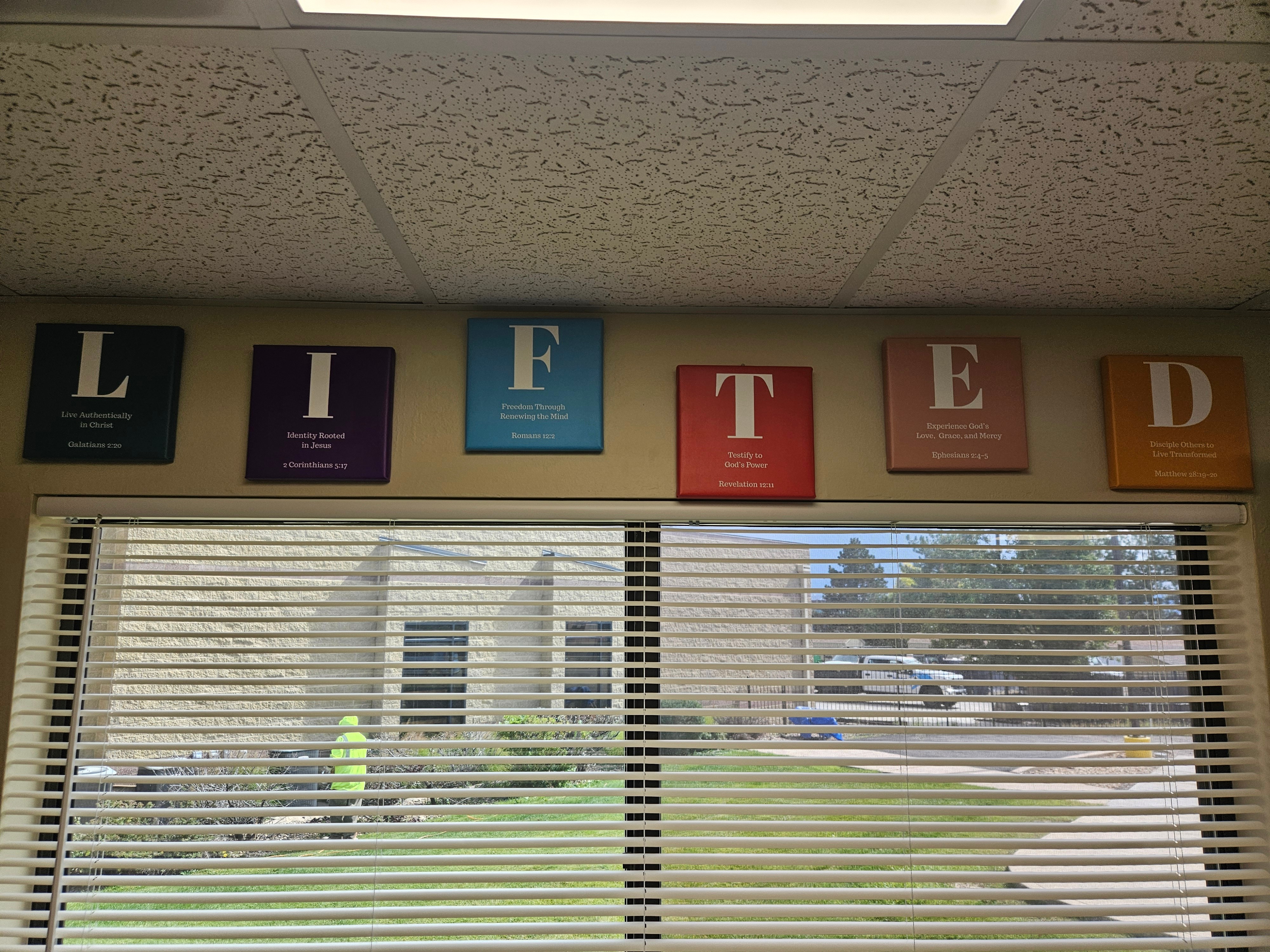 Six colorful signs with letters spelling 'LIFE' are on the wall above a window with blinds, showing a building and trees outside.