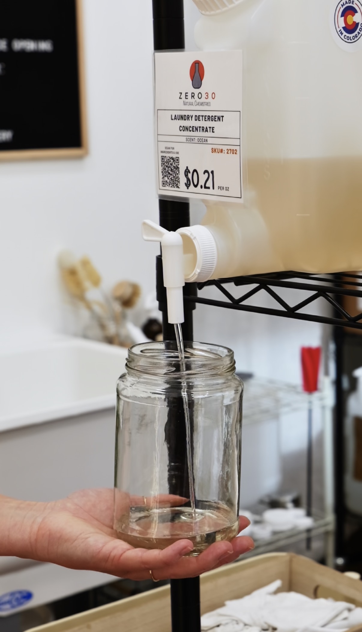 Person holding a glass jar under a dispenser with a white container above, in a laboratory setting.