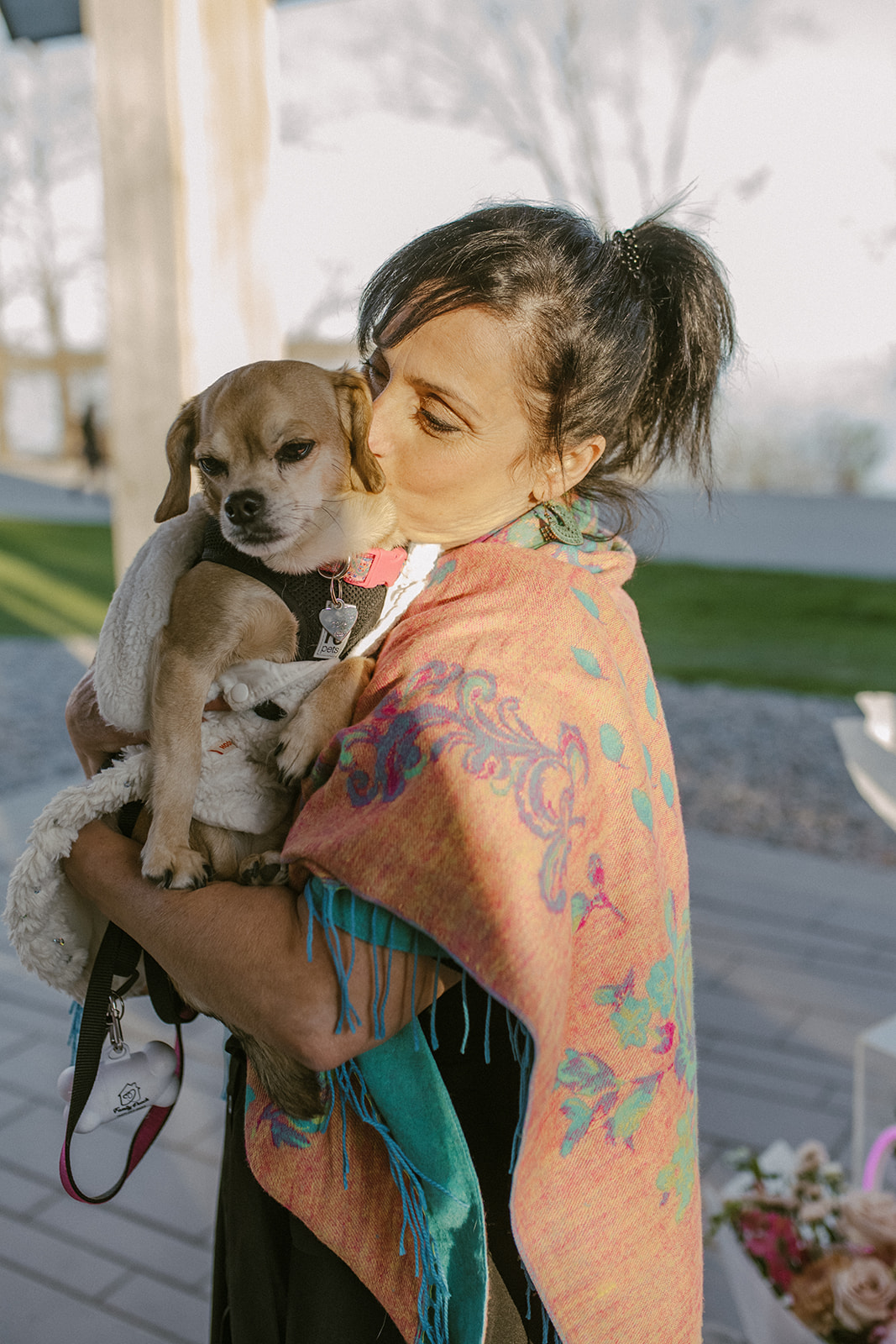 Woman holding a small dog outdoors, wearing a colorful shawl, with trees and a park in the background.