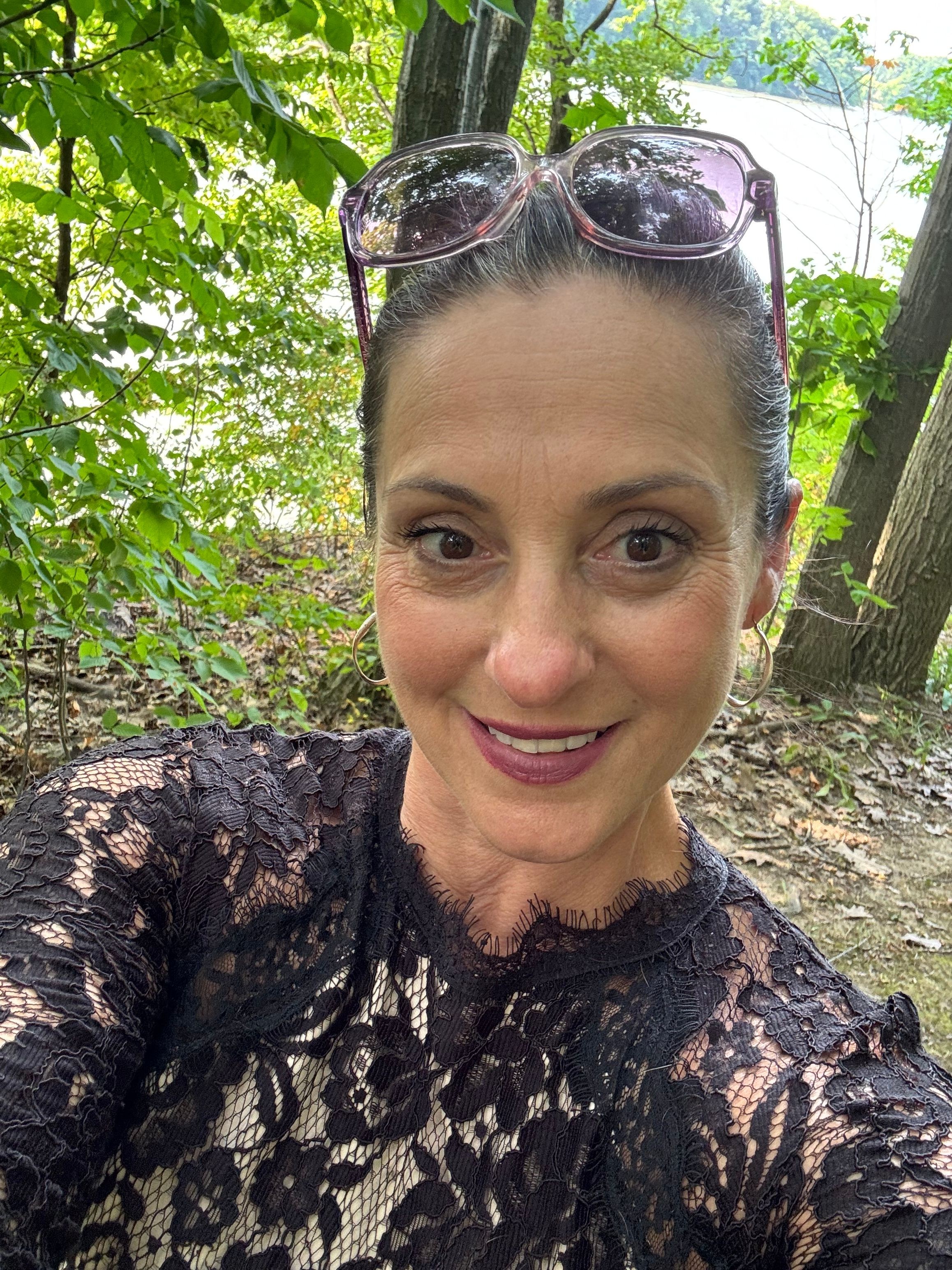 Woman with sunglasses on head, smiling, wearing black lace top, outdoors with green trees and sunlight.