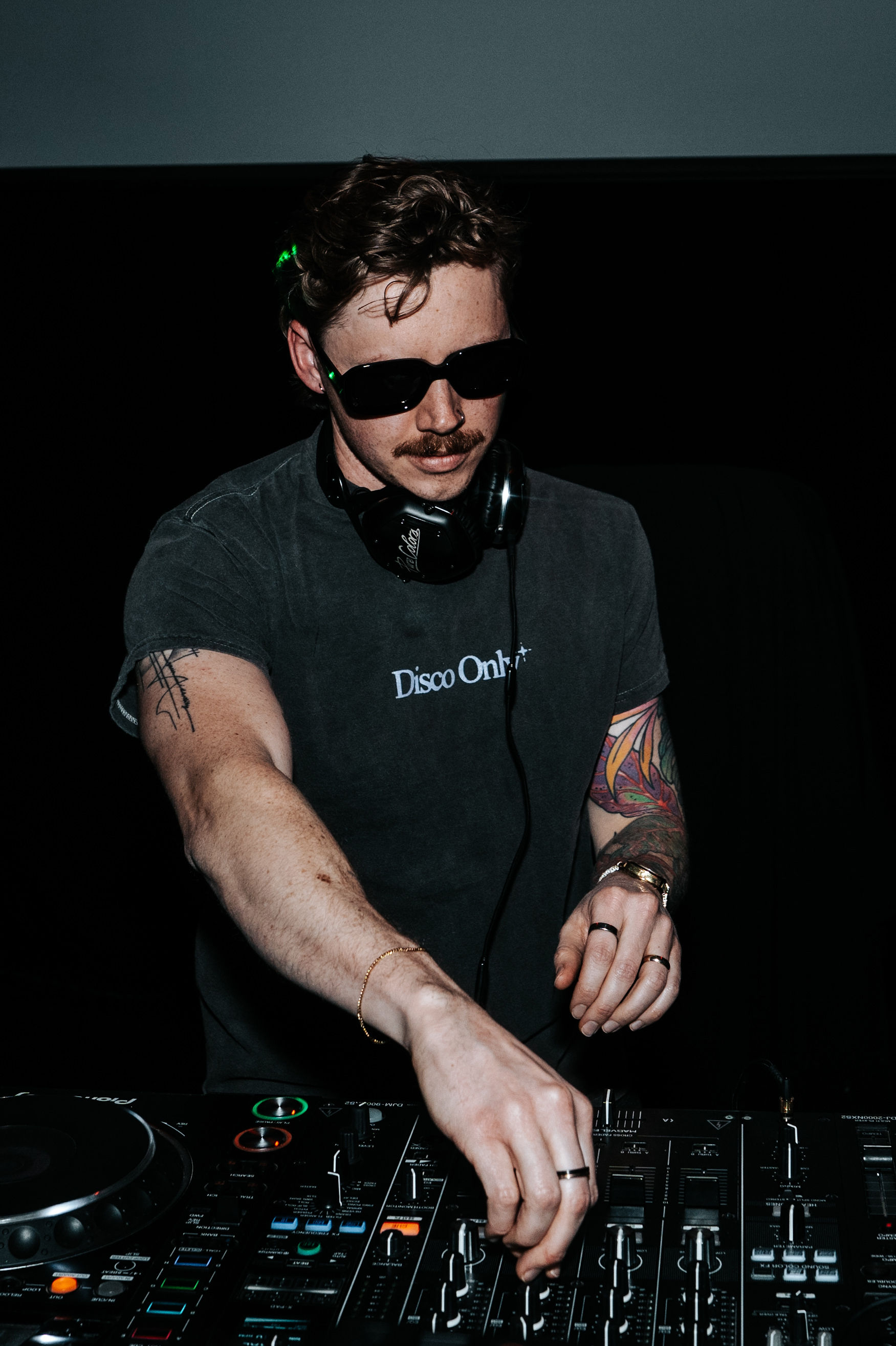DJ wearing sunglasses and headphones, operating a mixing console in a dark environment.