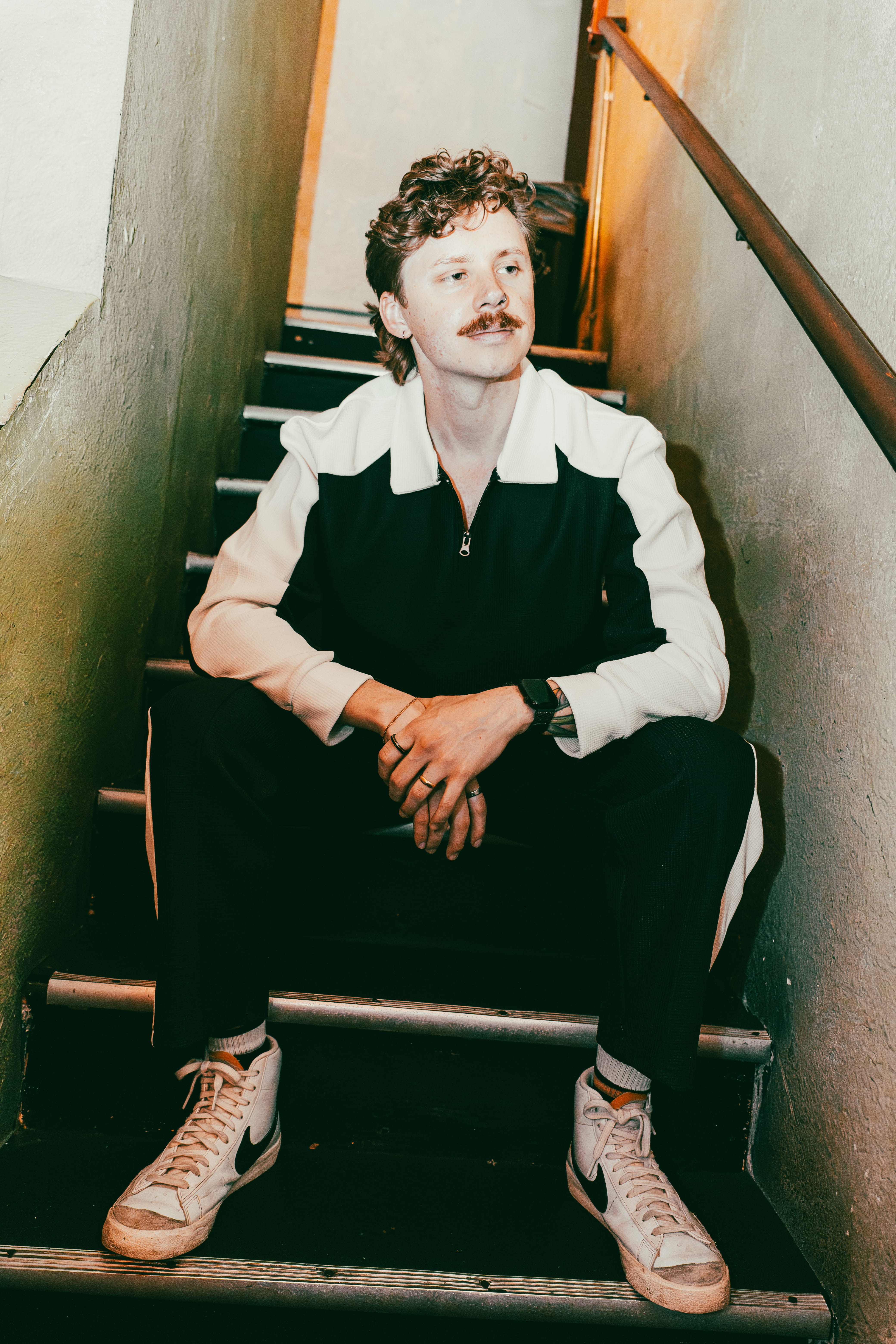 Person with curly hair and mustache sitting on stairs between narrow walls, wearing a black and white jacket and sneakers.