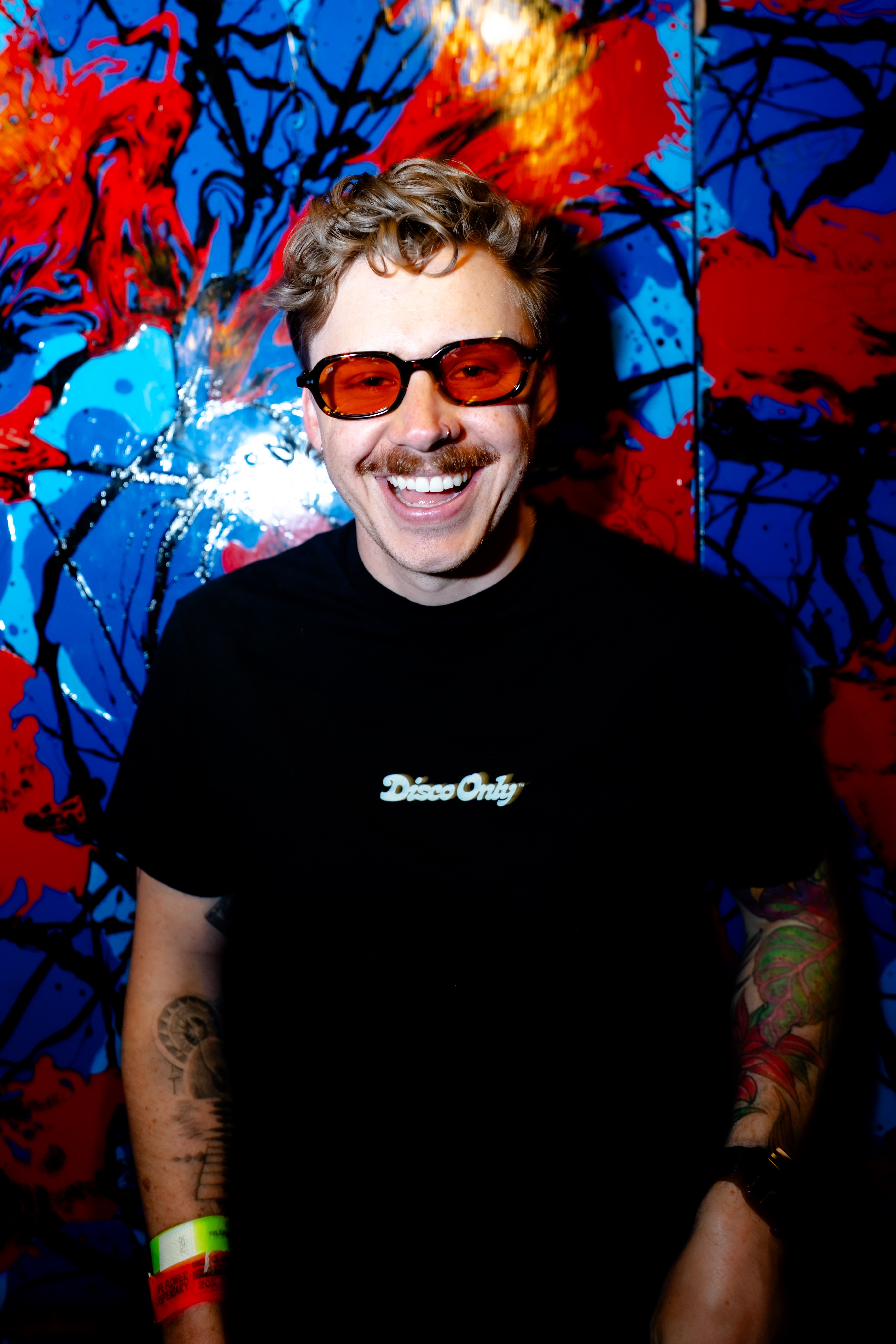 Person with curly hair, sunglasses, smiling, wearing a black shirt, standing in front of colorful abstract background.