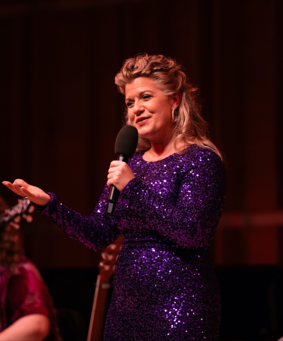 Woman in a sparkly purple dress speaking into a microphone on stage, gesturing with her hand.