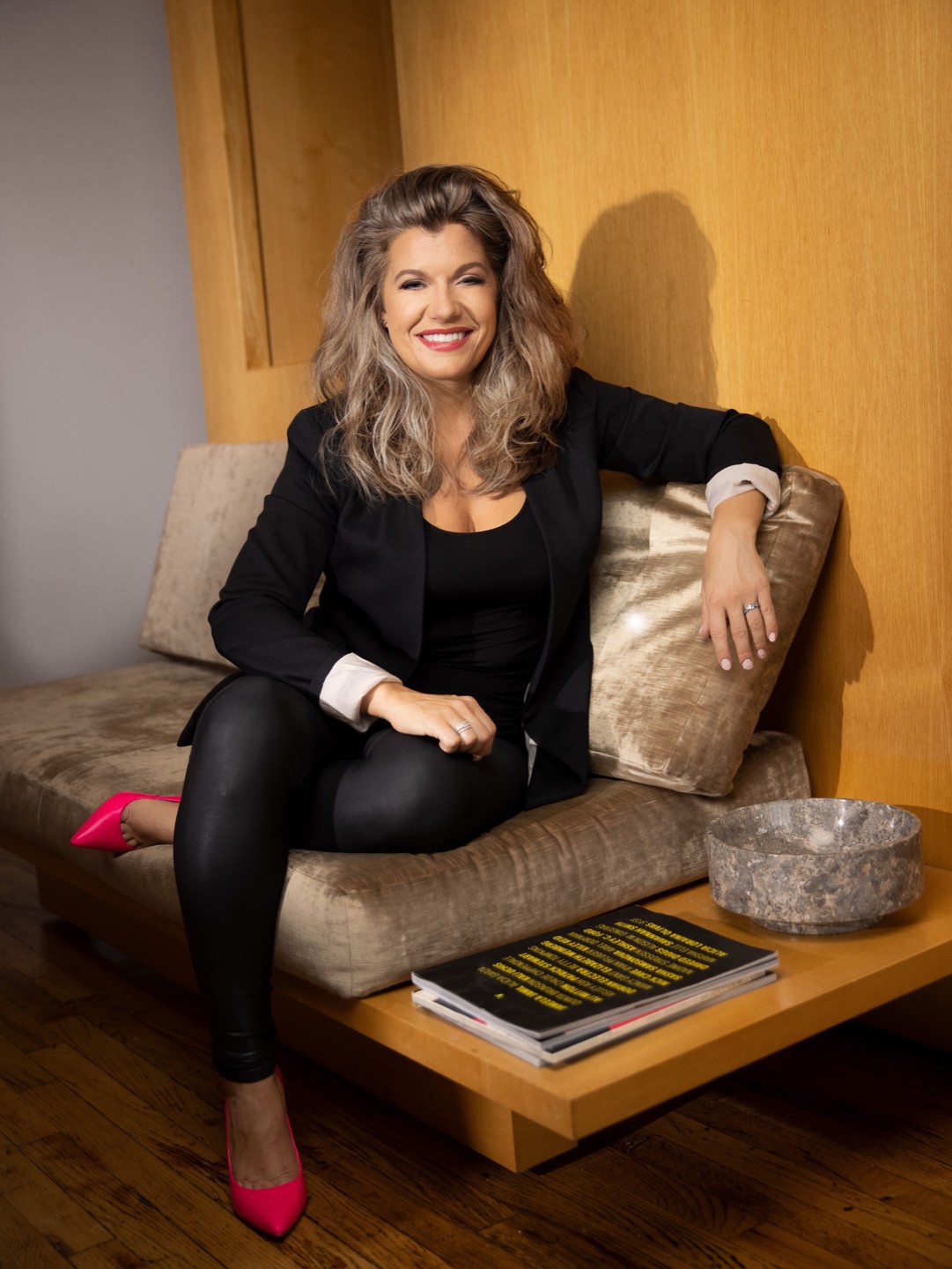 Woman with wavy hair sitting on a sofa, smiling, wearing a black outfit and pink shoes, in a room with wooden wall.