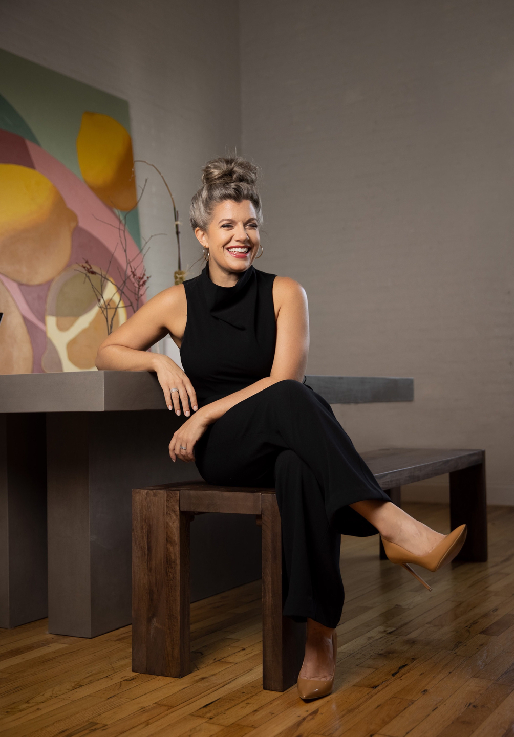 Woman with gray hair in a bun, smiling, sitting on a bench in a room with abstract art, wearing a black sleeveless top and pants.