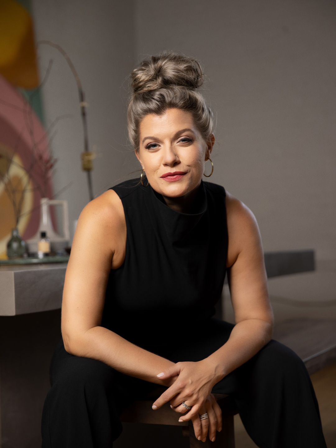Woman with styled hair and hoop earrings sitting indoors, wearing a black sleeveless top, looking at the camera.