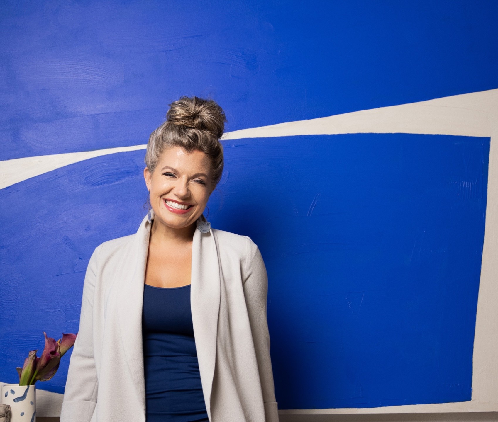 Smiling woman with a bun hairstyle standing in front of a blue wall with white abstract design, wearing a white blazer and navy top.