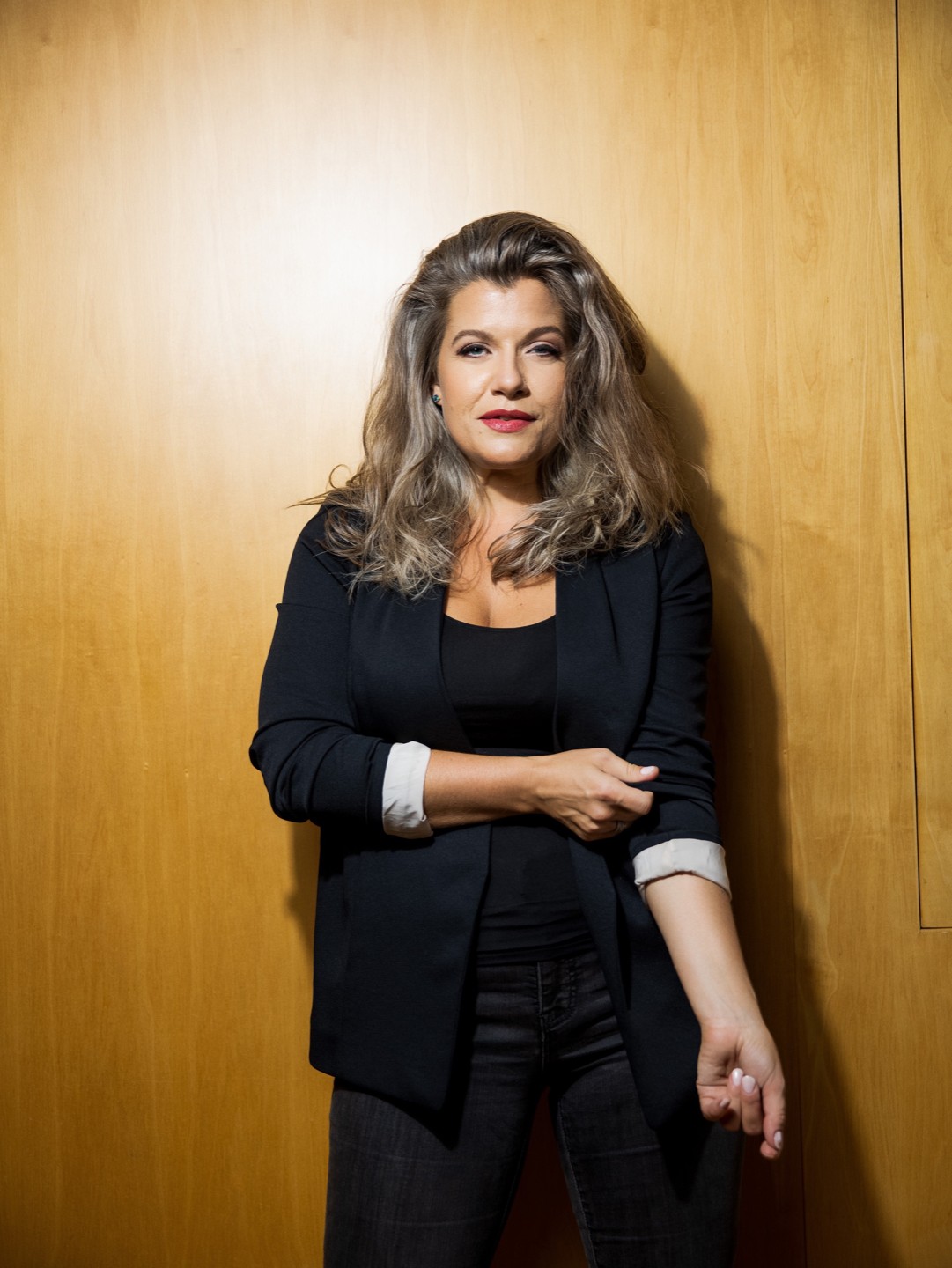 Woman with wavy hair wearing a black blazer and pants, standing against a wooden wall.
