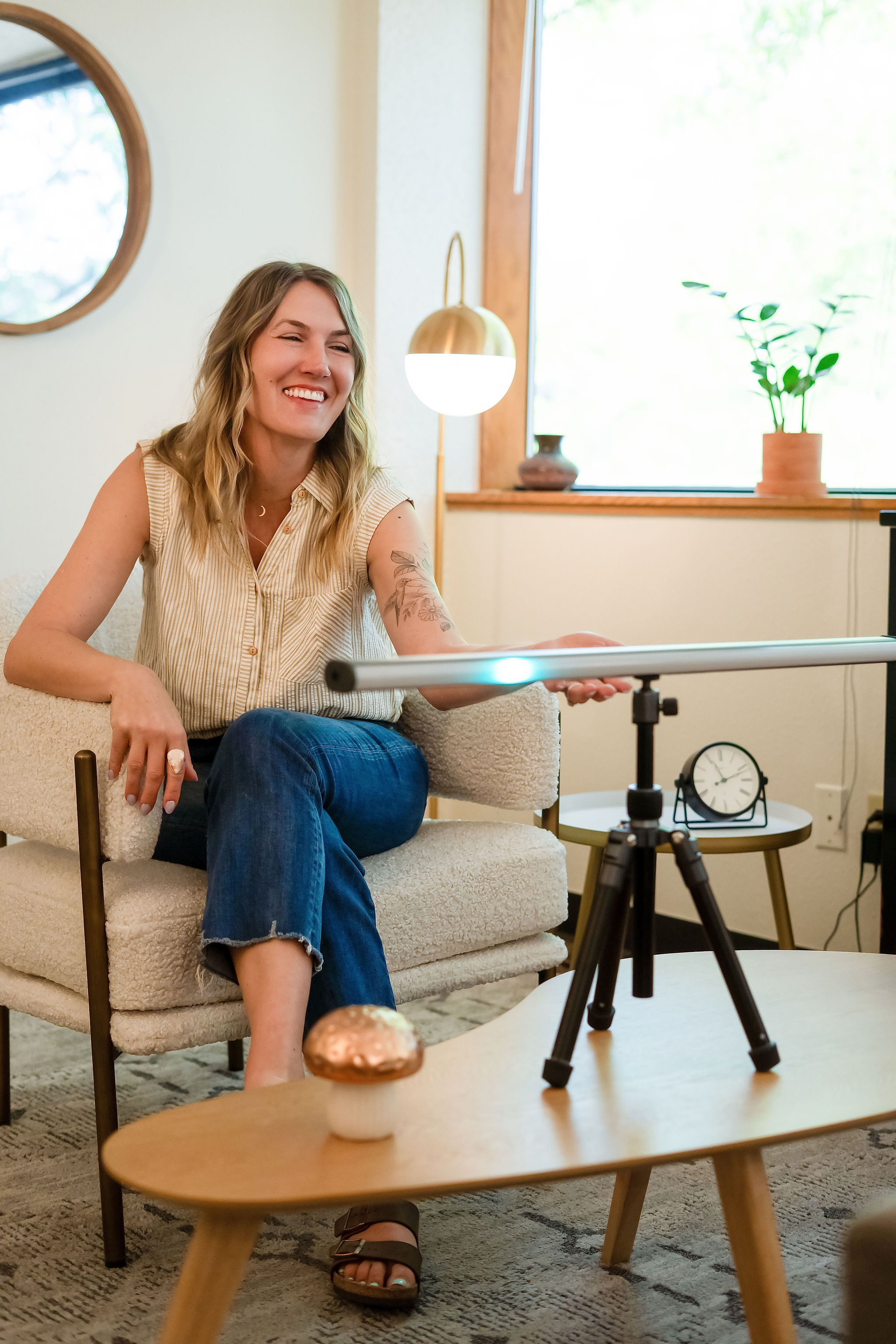 Woman sitting on a sofa, smiling, with a camera on a tripod in front of her, in a bright room with a window.