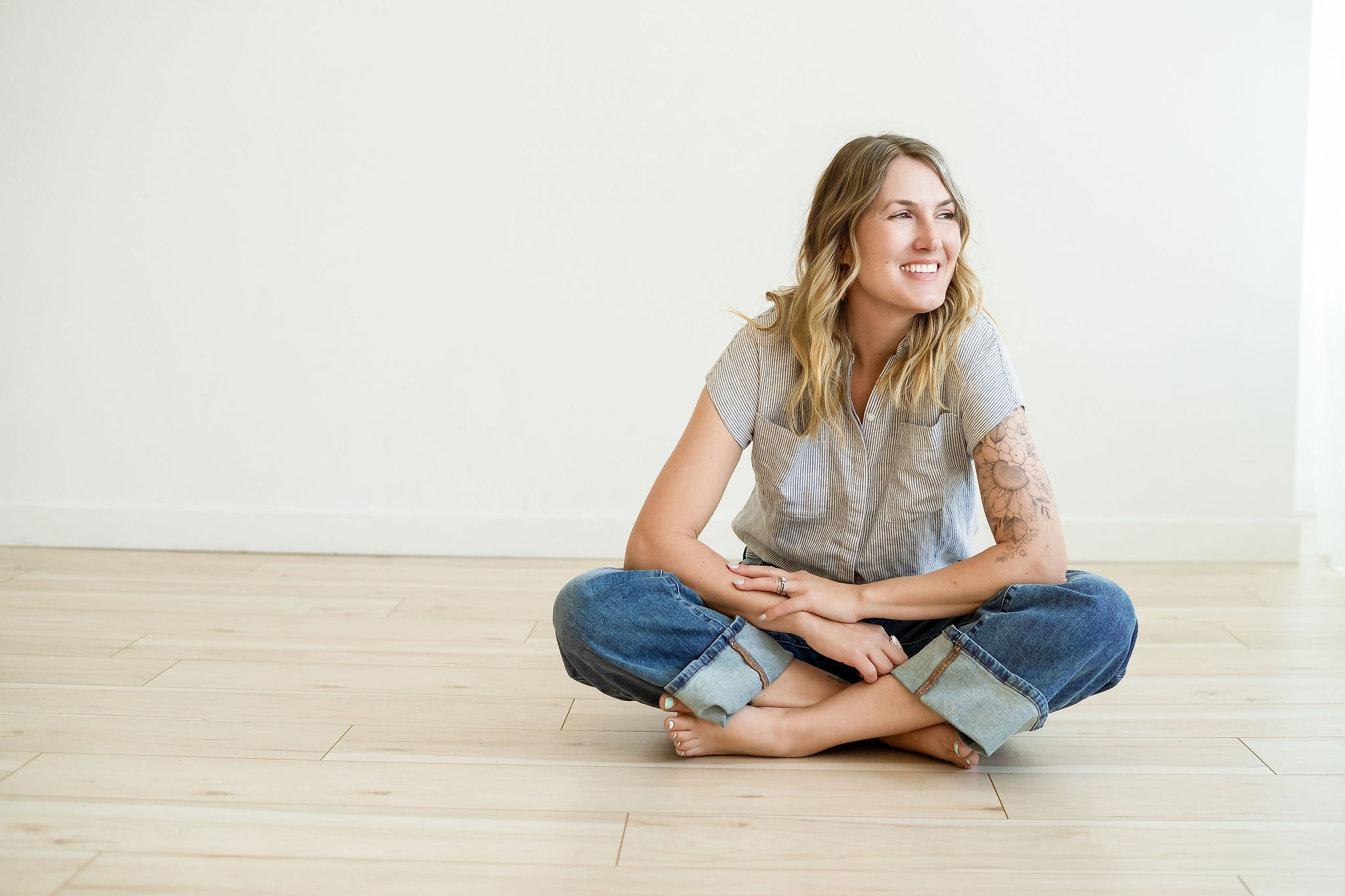 Young woman sitting cross-legged on a light wooden floor, smiling, with a plain white wall behind her.