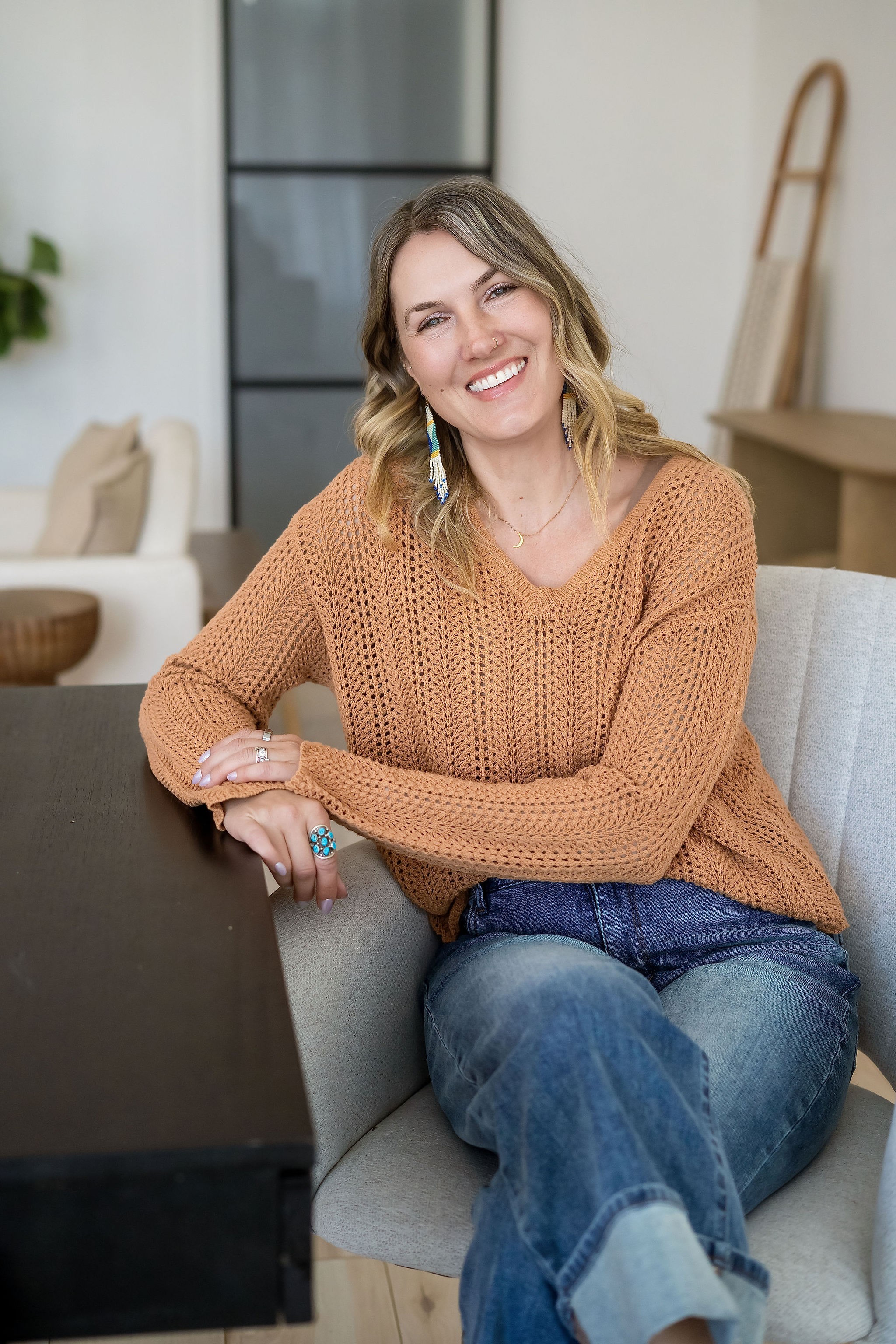 Woman with shoulder-length hair smiling, sitting on a sofa in a cozy room, wearing a brown sweater and blue jeans.