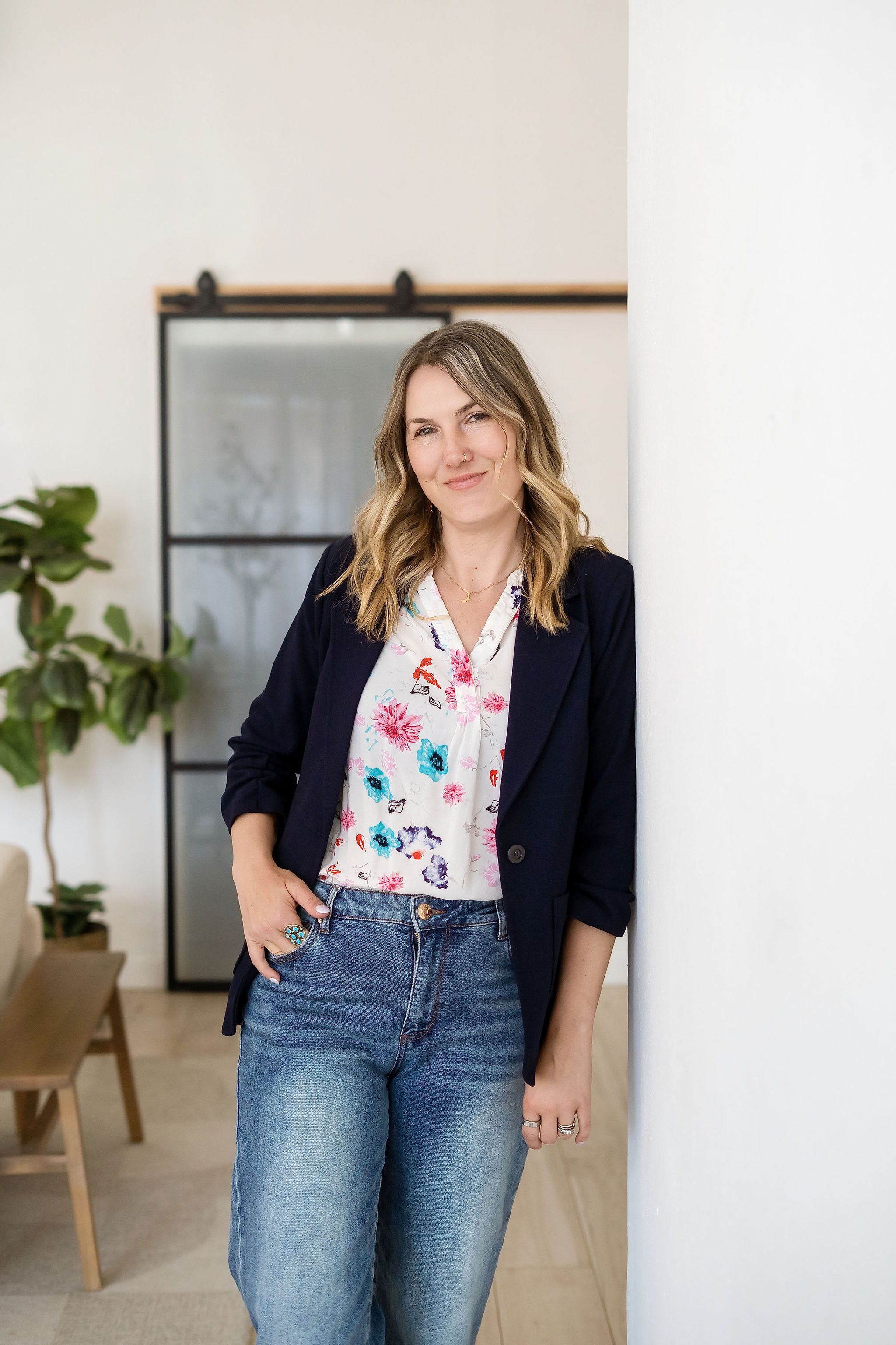 Woman with shoulder-length hair wearing a floral shirt and dark blazer, standing indoors near a white wall.
