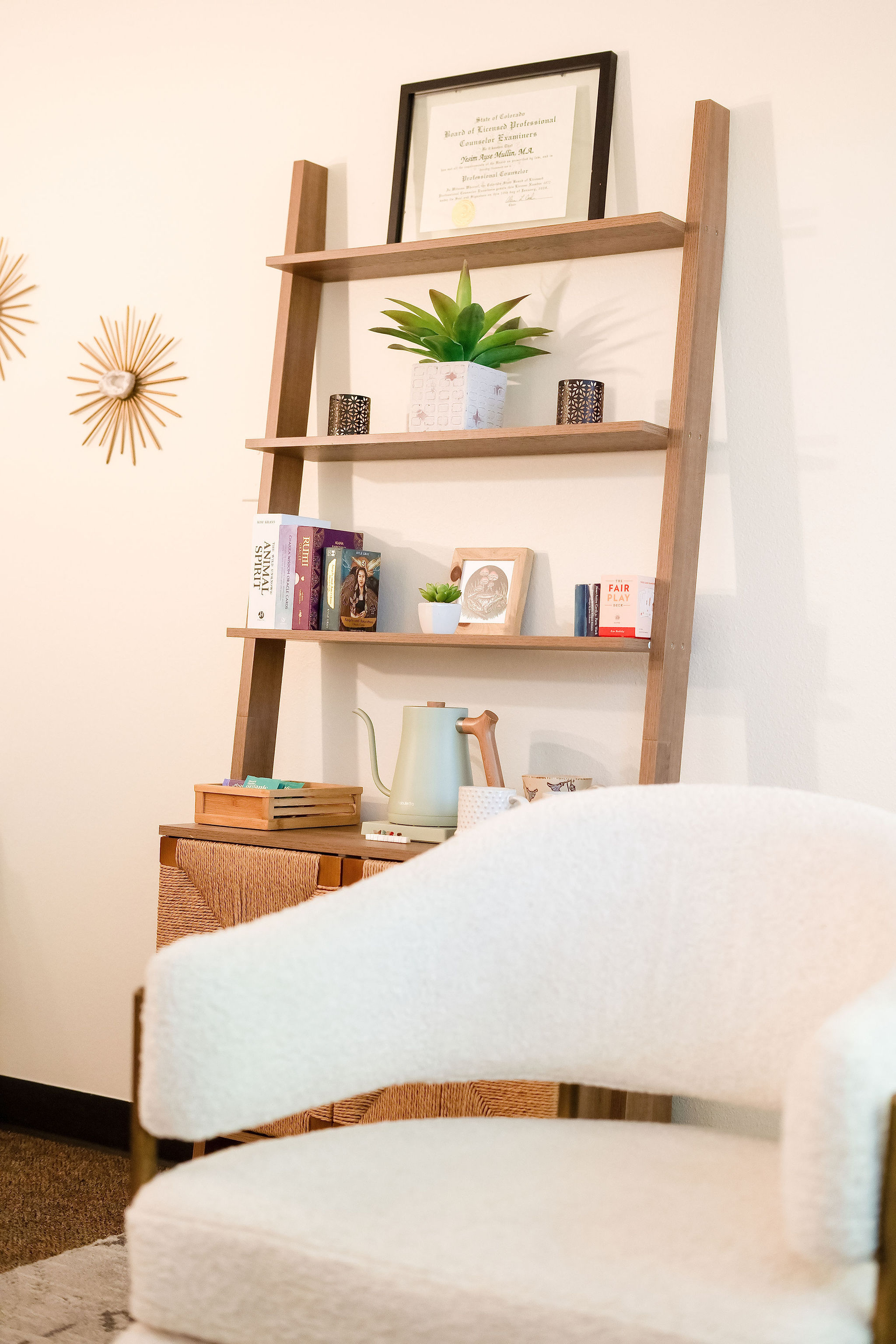 Wooden ladder-style shelf with books, a potted plant, and decorative items, next to a white armchair and wall decor.