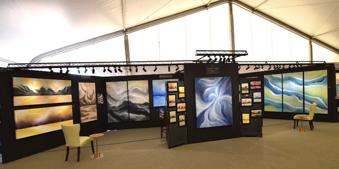 Art exhibition with landscape and abstract paintings displayed on walls and panels, chairs in front of artwork, well-lit space.