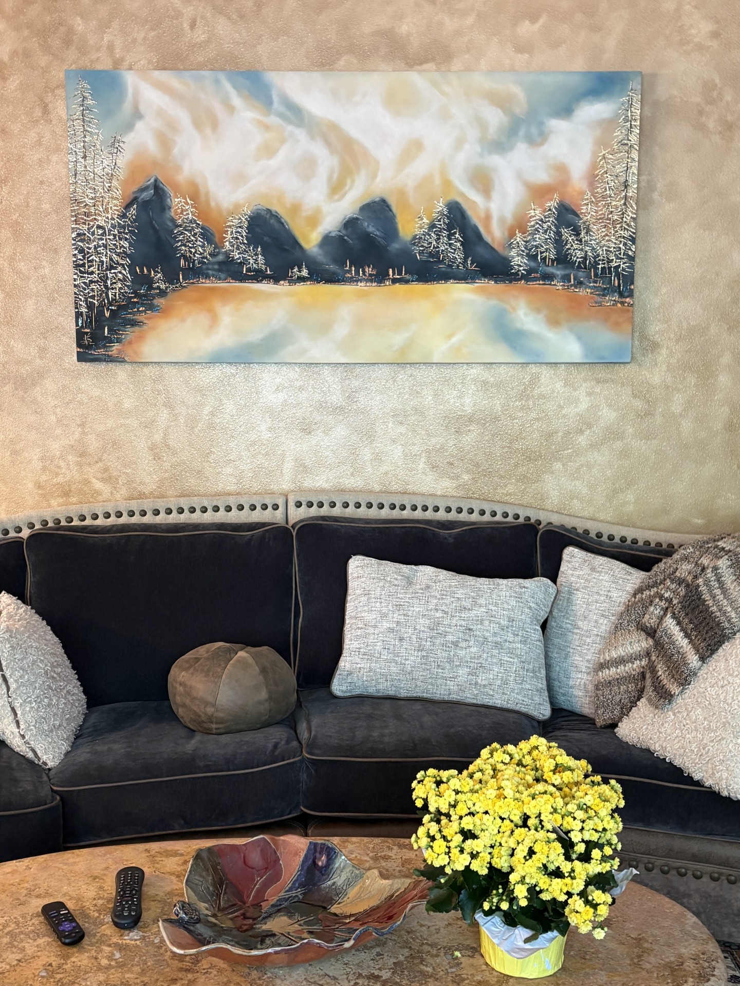 Painting of mountains and trees above a lake hangs on wall, black sofa with pillows, yellow flowers on table.