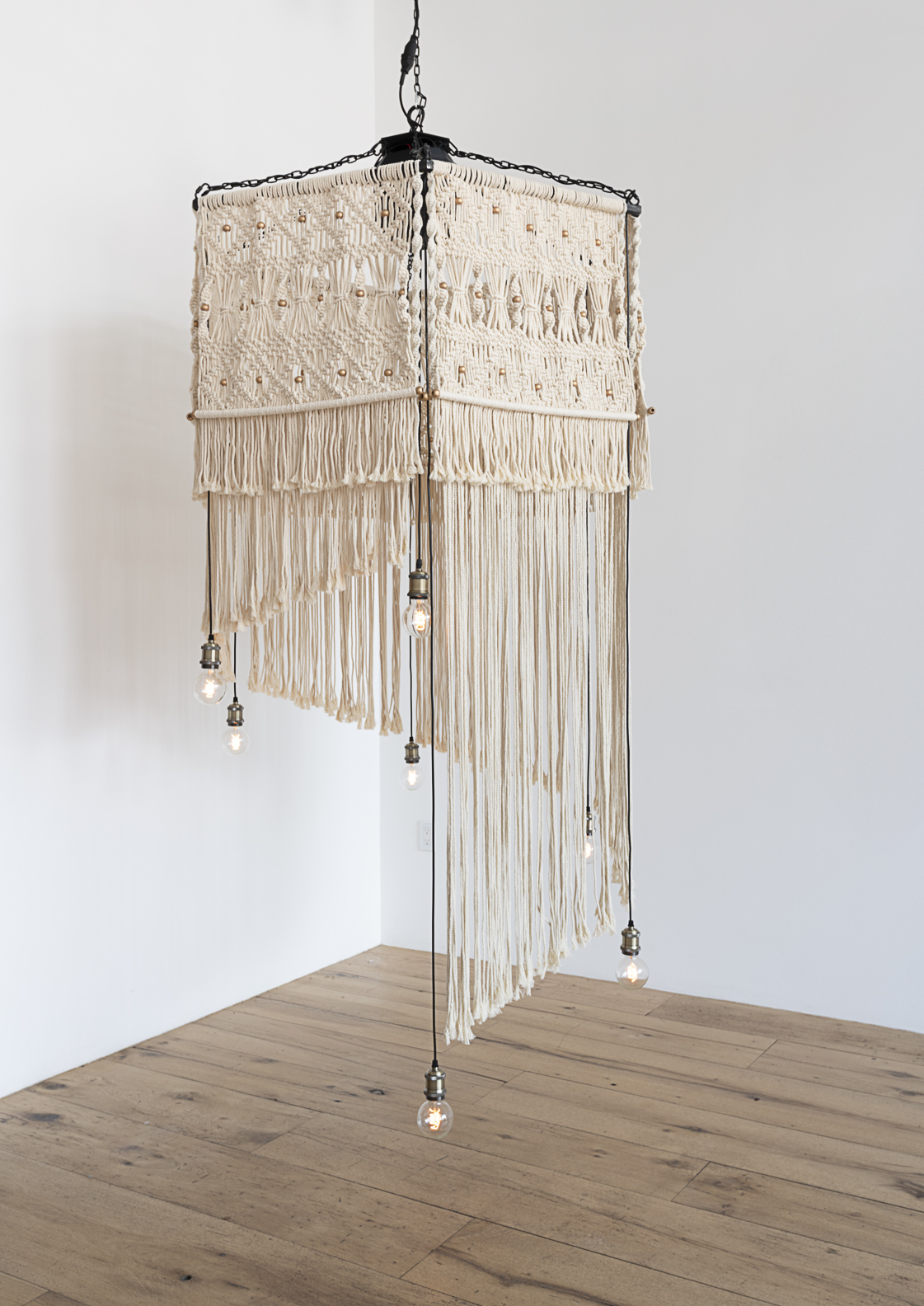 Decorative hanging lamp with fabric and fringe, suspended from ceiling, against plain white wall and wooden floor.