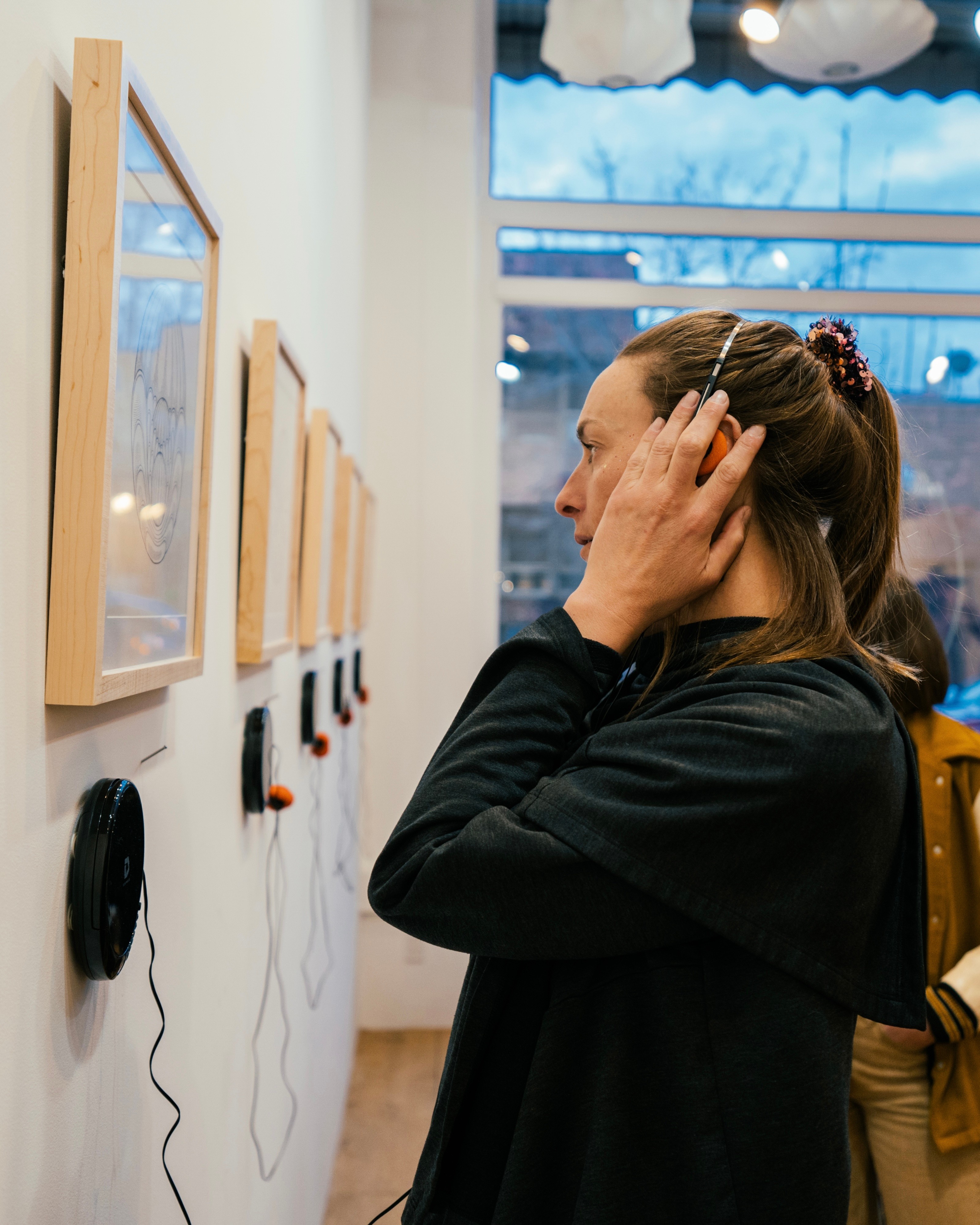 Woman with hair in a ponytail looking at artwork on a wall, touching her ear, in a gallery setting.