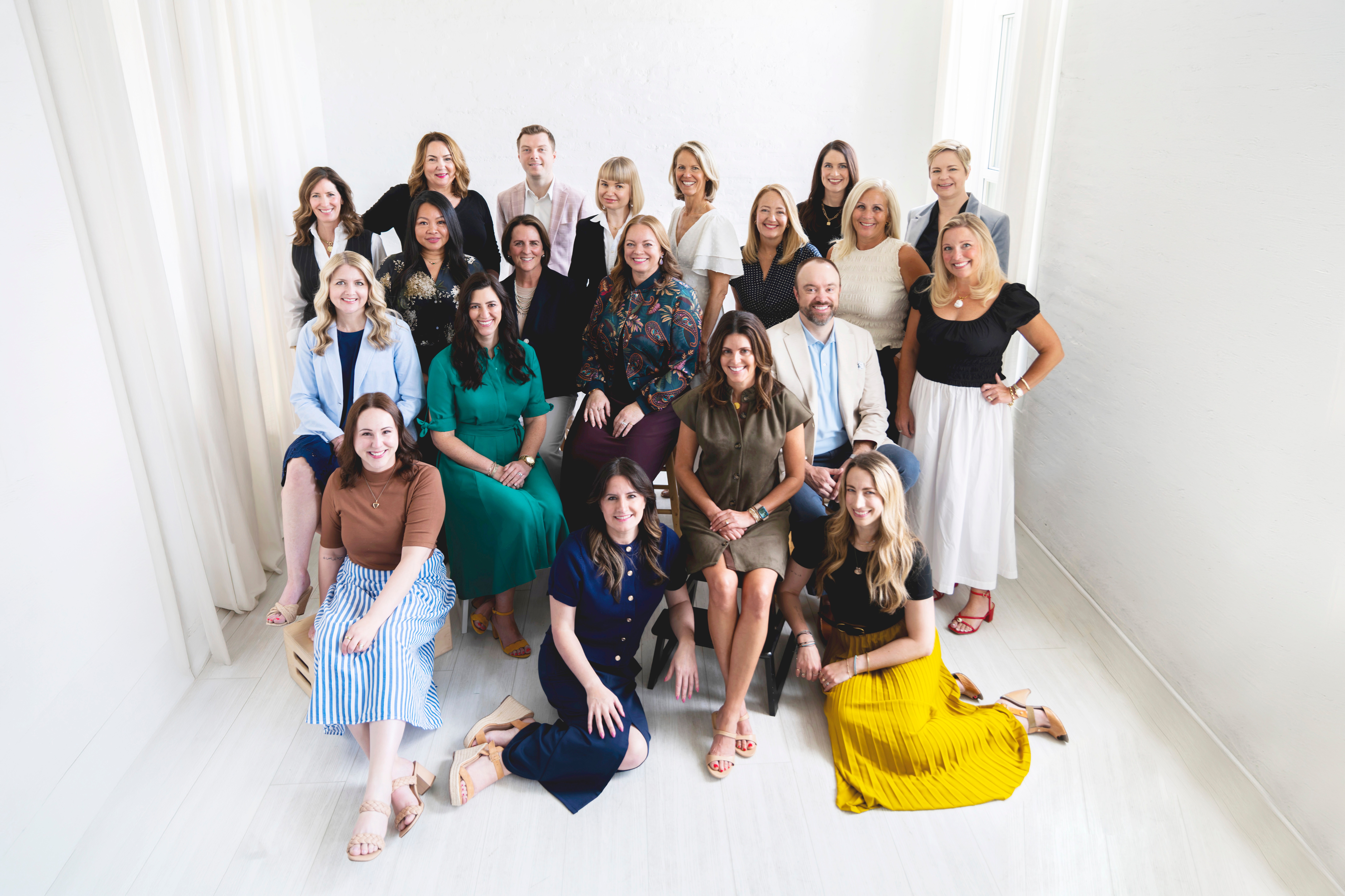 Group of 20 women and girls posing in a bright, white room with sheer curtains, some sitting and some standing.