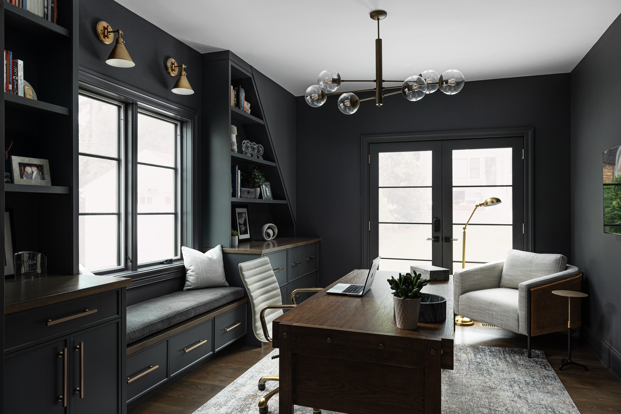 Living room with dark walls, large windows, a wooden table, a white armchair, and a built-in bookshelf, with a door leading outside.