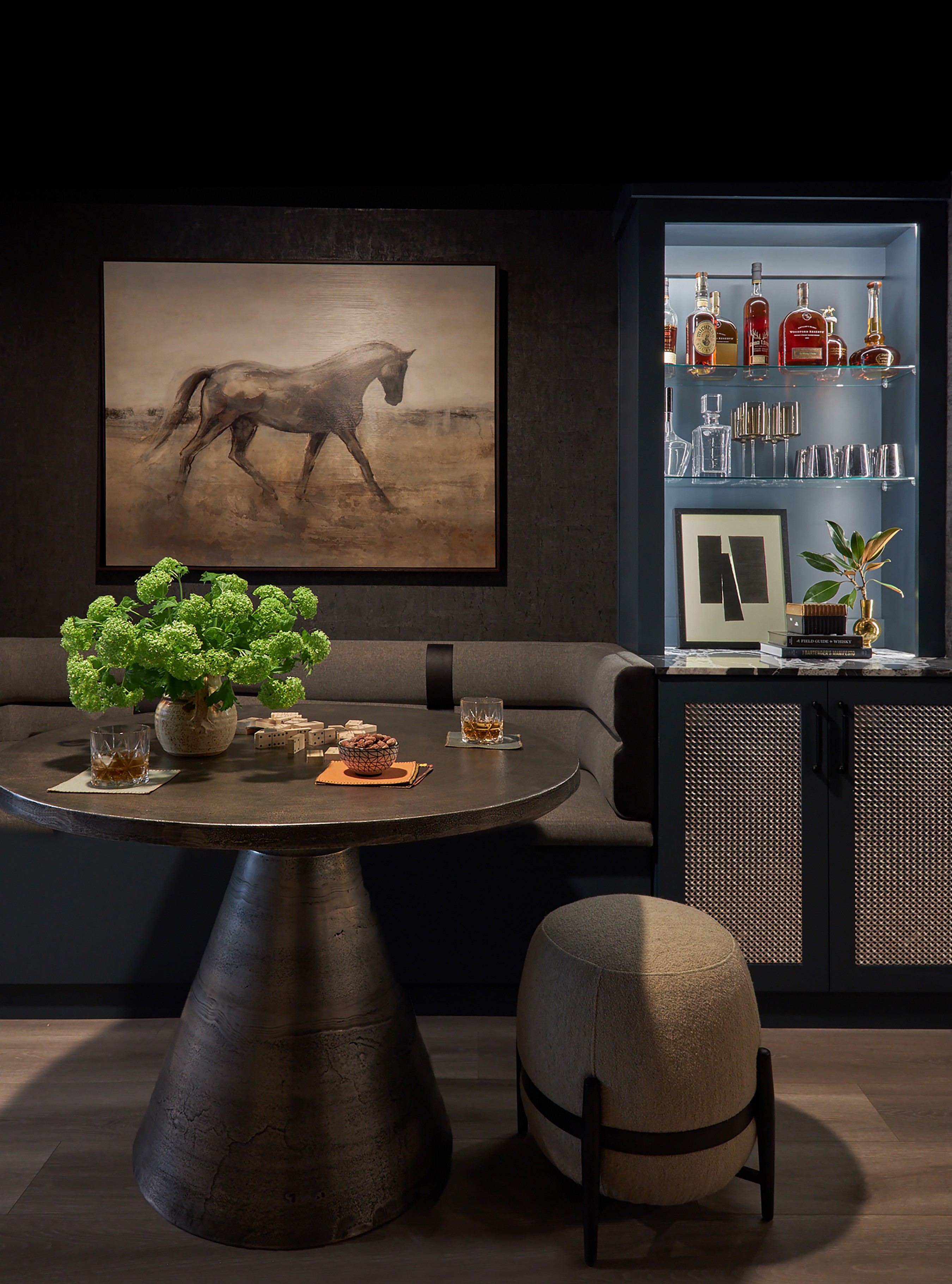 Living room with a round table, a beige stool, a painting of a horse, and a cabinet with bottles and glasses.