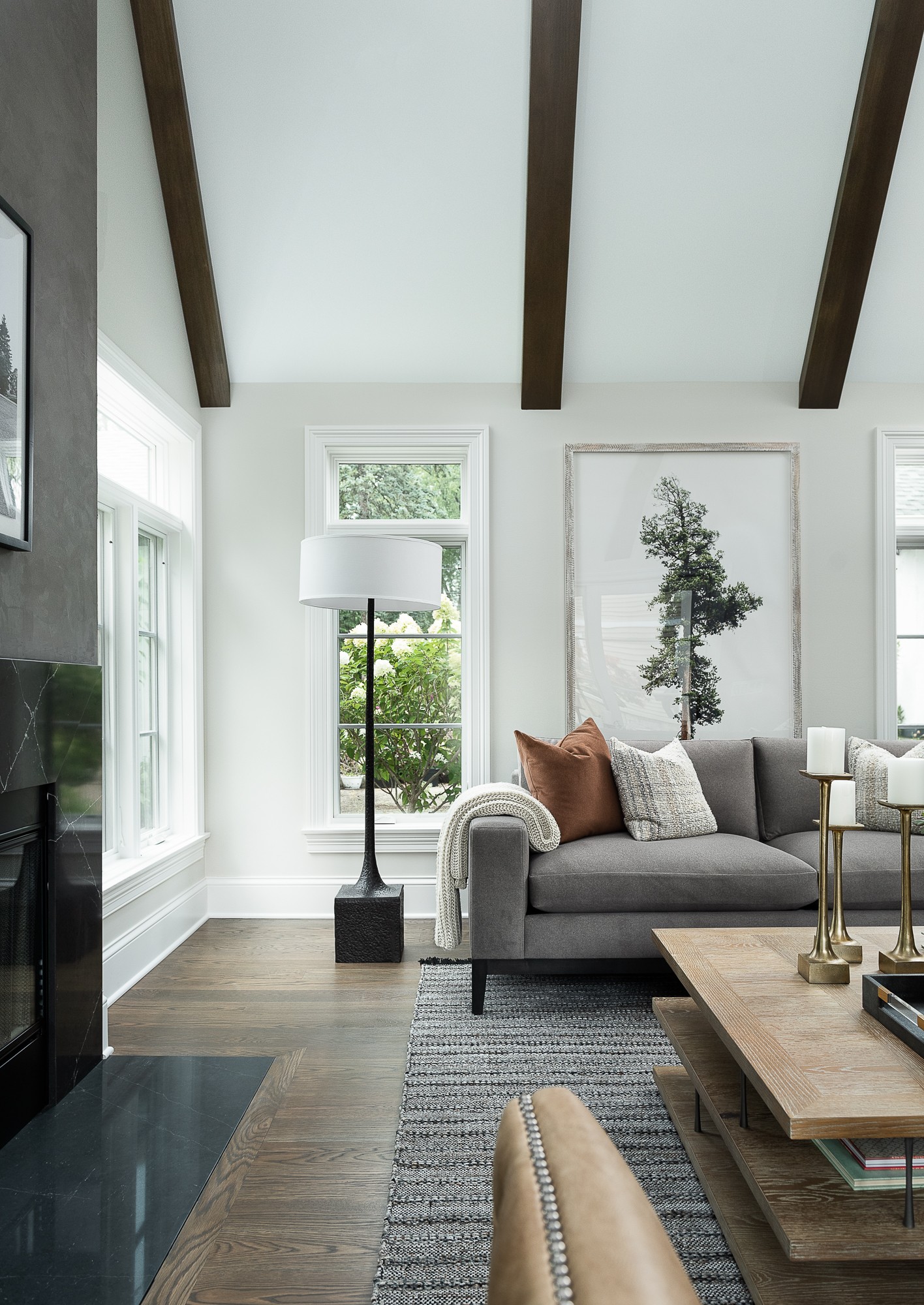 Living room with white walls, large windows, gray sofa with pillows, wooden ceiling beams, and a wooden coffee table.