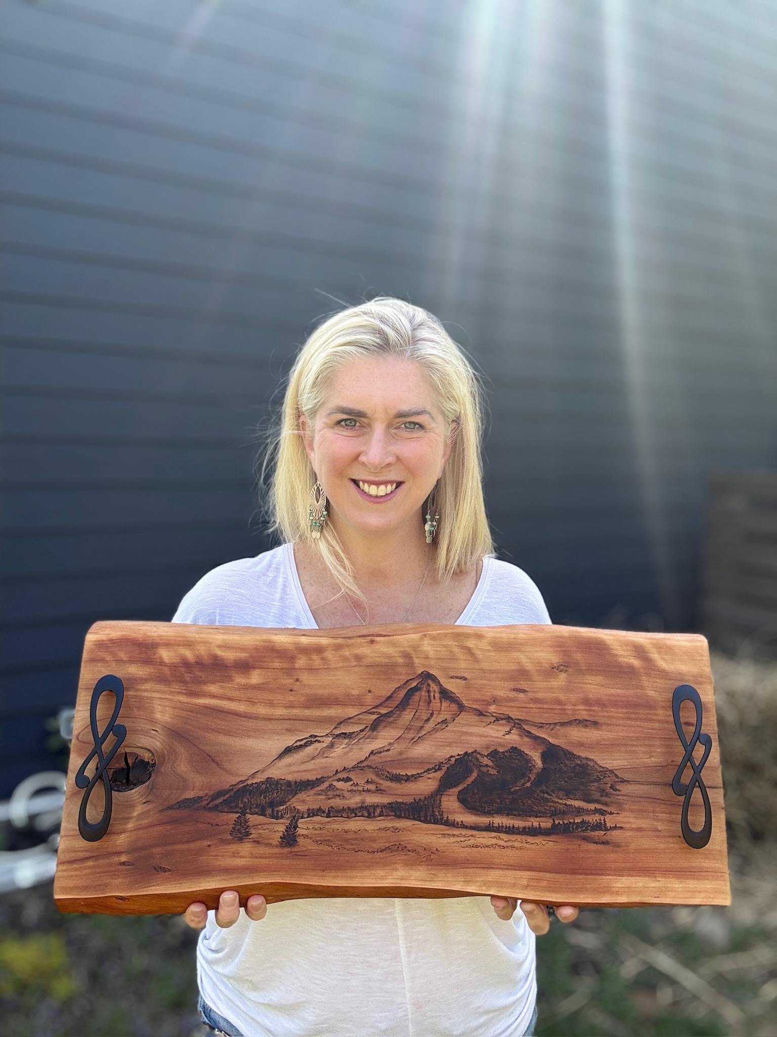 Woman with blonde hair smiling holding a wooden artwork of a mountain landscape outdoors.