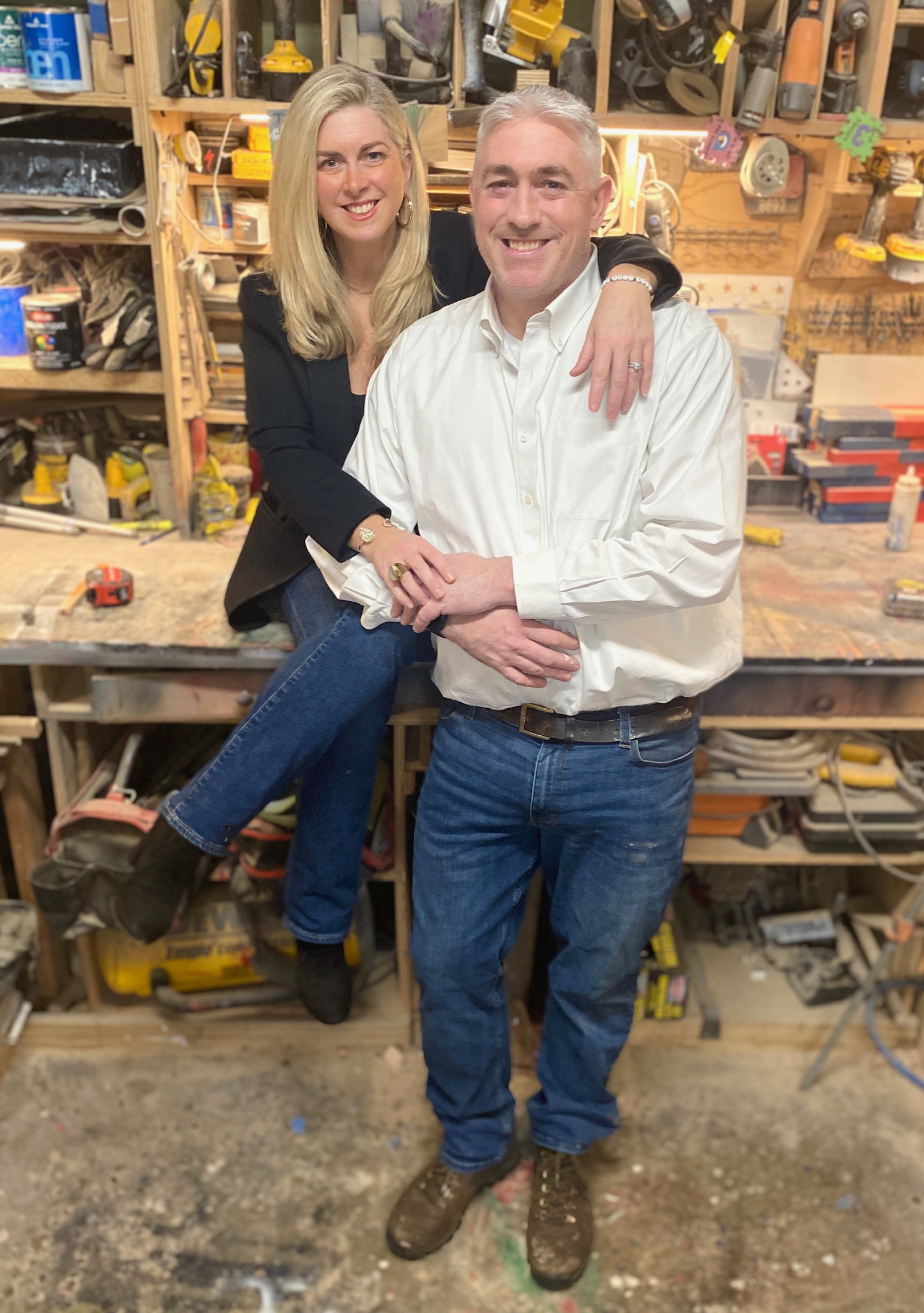 A smiling man and woman pose together in a workshop, with the woman sitting on a workbench and the man standing, both smiling.