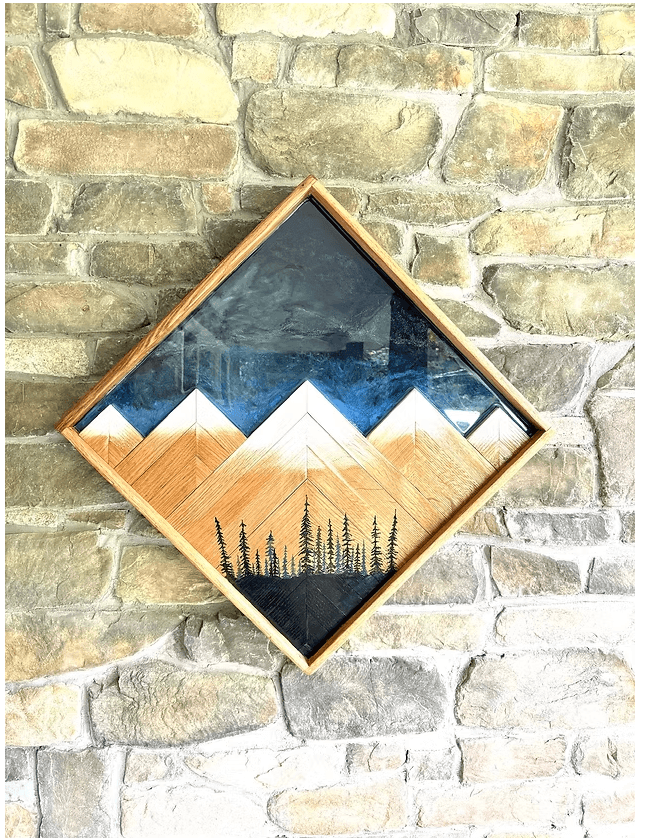 Framed mountain landscape with trees, hanging on a brick wall, in a diamond orientation.