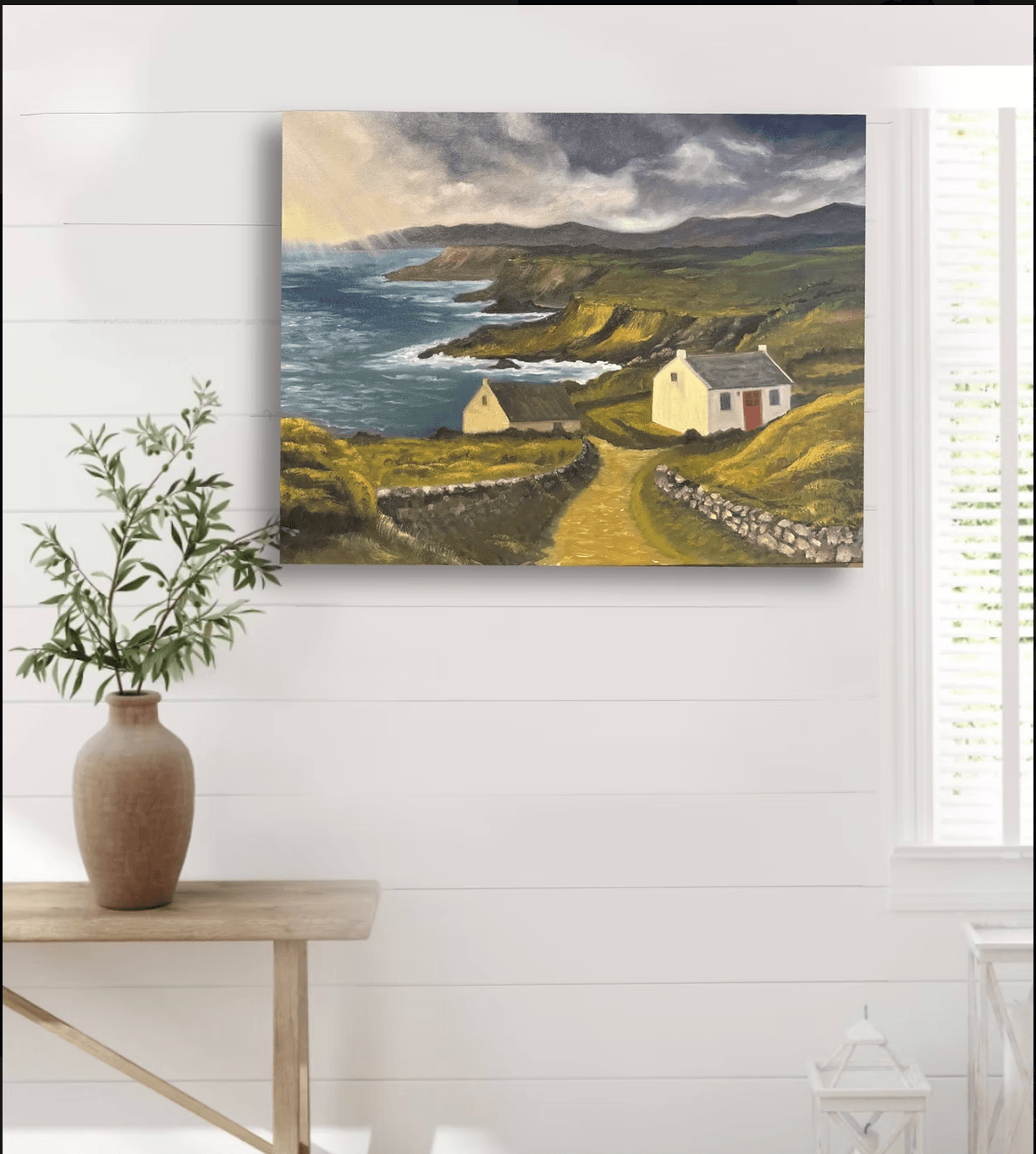 Painting of a coastal landscape with houses, cliffs, and a cloudy sky, hanging on a white wall.