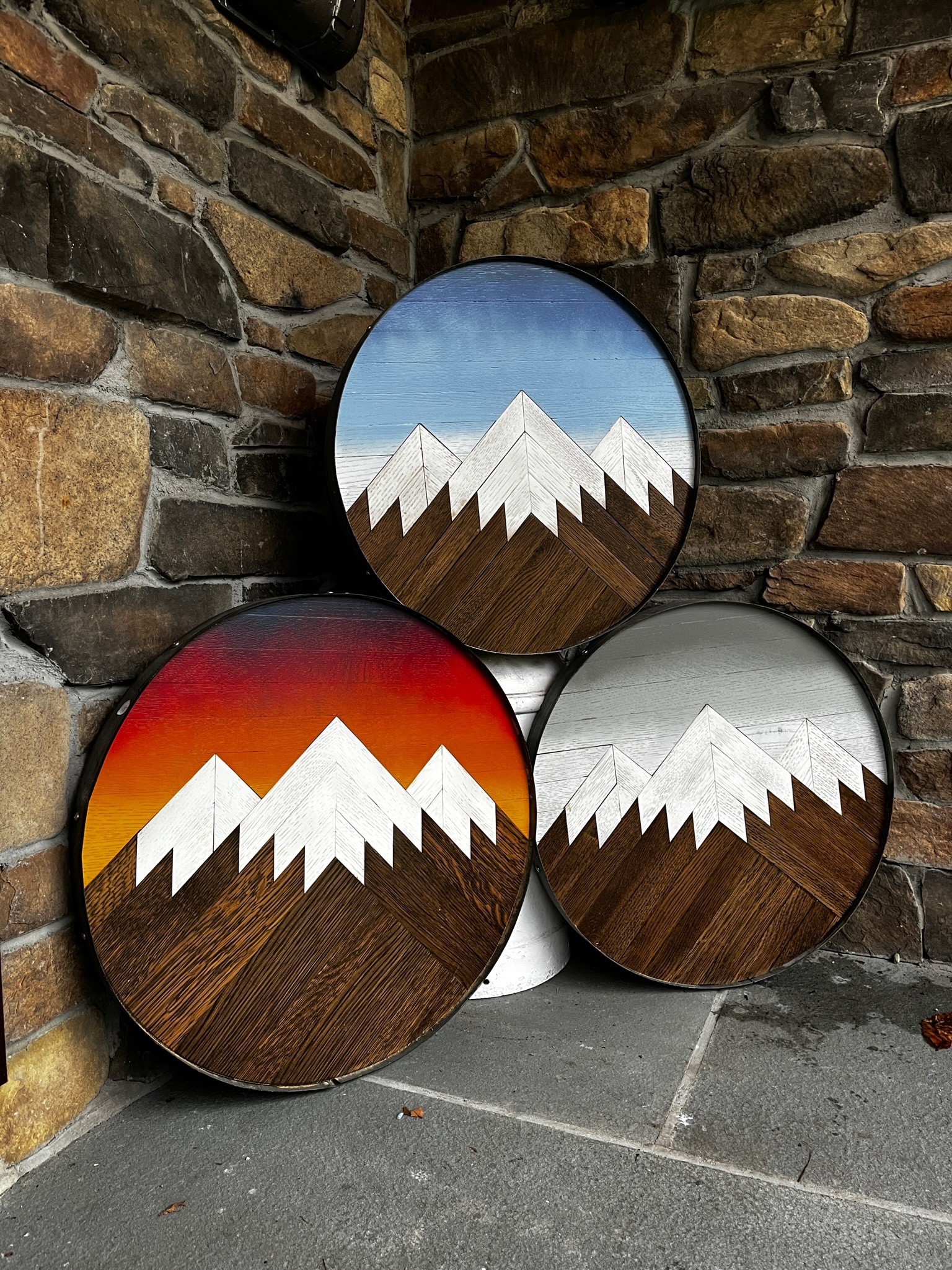 Four circular wall decorations with mountain designs, set against a brick wall. Each features a different sky background: blue, sunset, and gray.