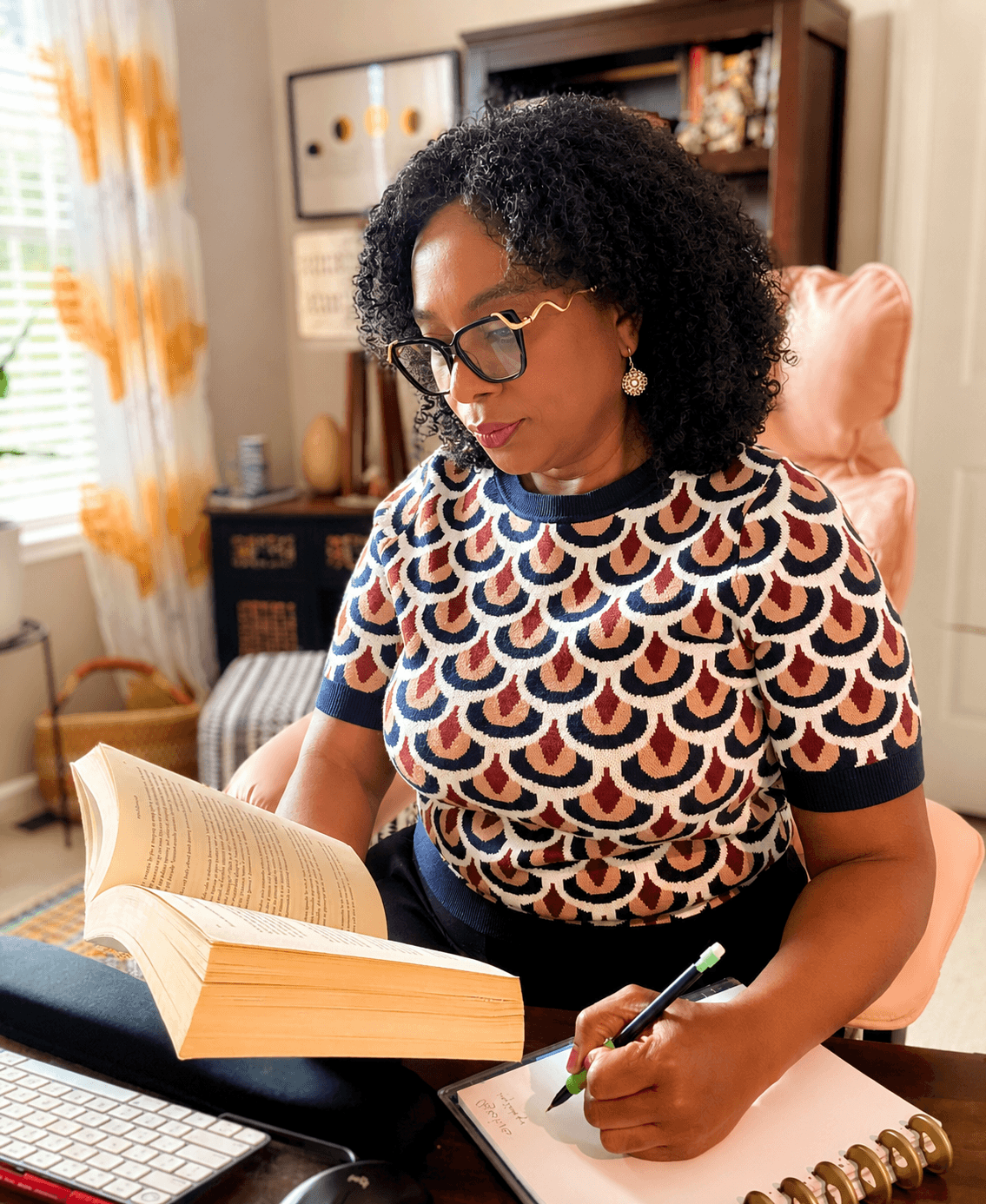 Woman with curly hair and glasses reading a book at a desk, writing on paper, in a cozy room.