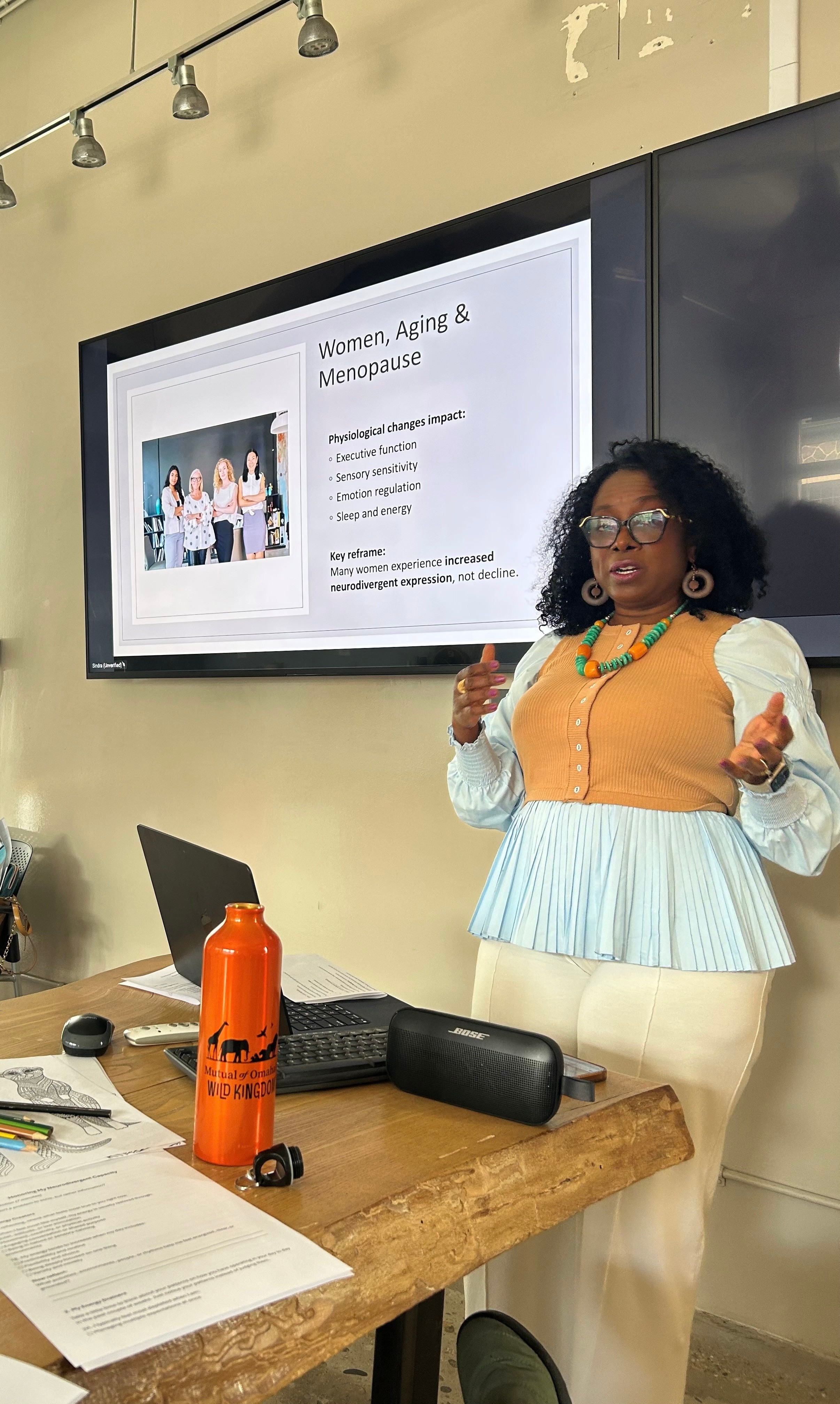 Woman giving presentation in a classroom, standing next to a screen with a slide about women, aging, and menopause.